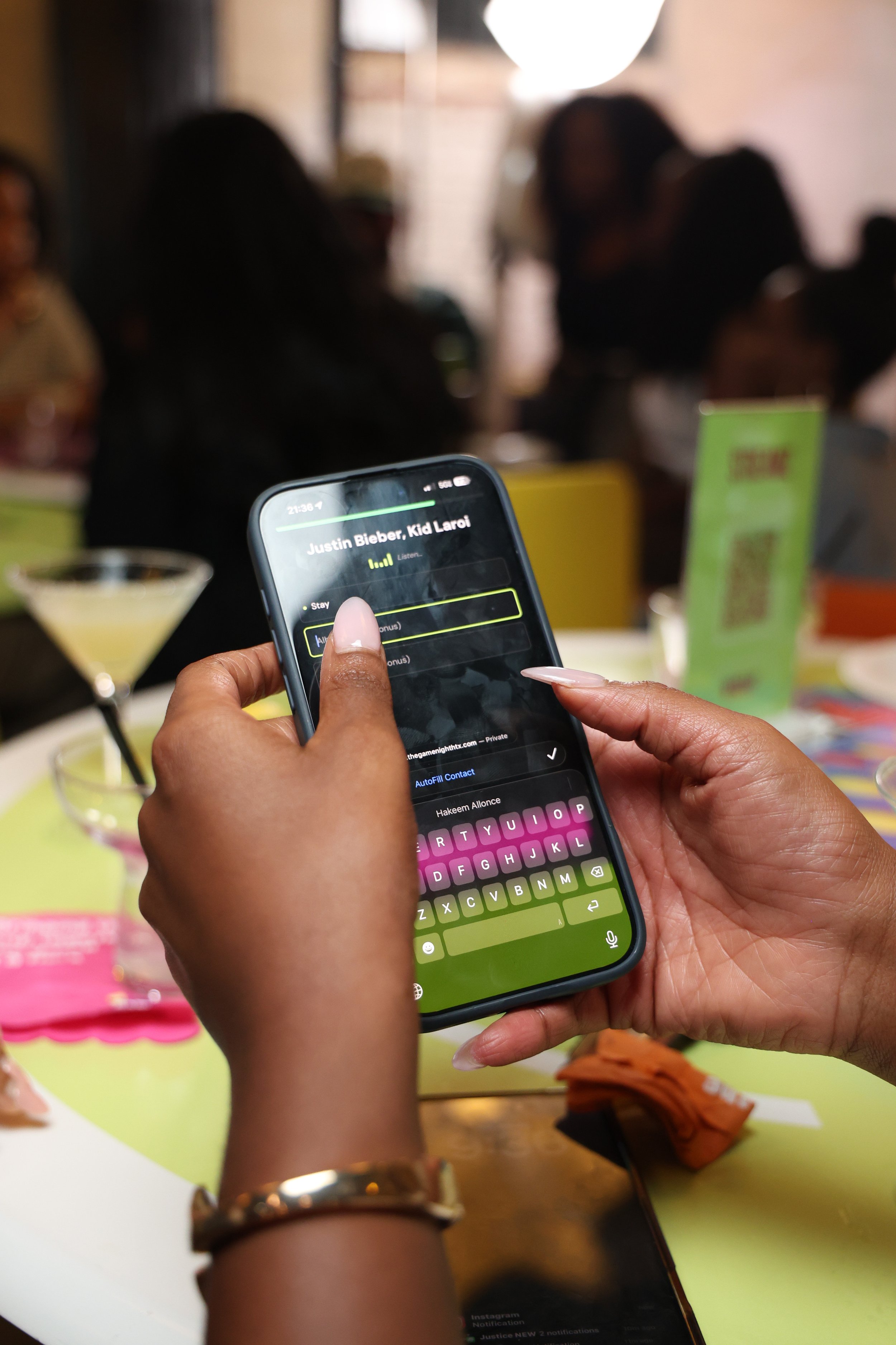Person holding a smartphone with a music player app open, playing Justin Bieber's 'Kid Laori' in a lively indoor setting with blurred people in the background.
