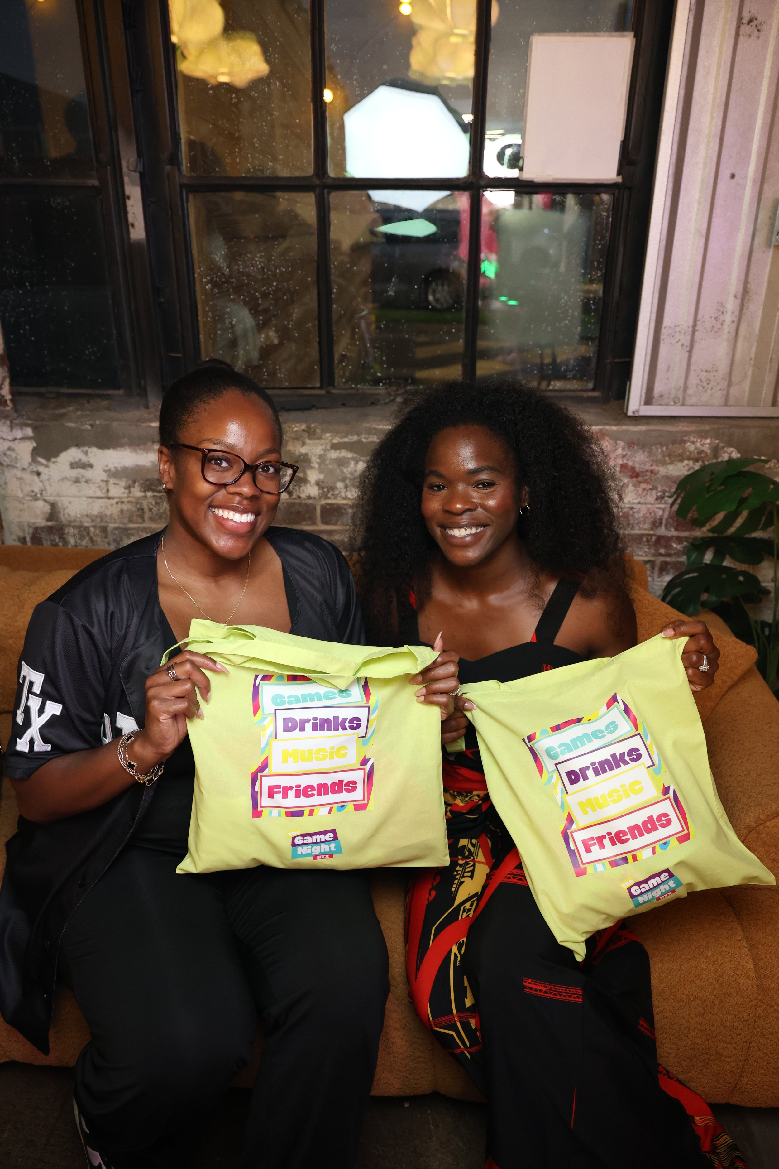 Two women sitting on a brown couch inside a restaurant, holding yellow tote bags that read "Games, Drinks, Music, Friends. Game Night." The woman on the left wears glasses, a black jacket, and jewelry; the woman on the right has curly hair and a black top with red and yellow accents. Behind them is a large window with raindrops, and a brick wall.