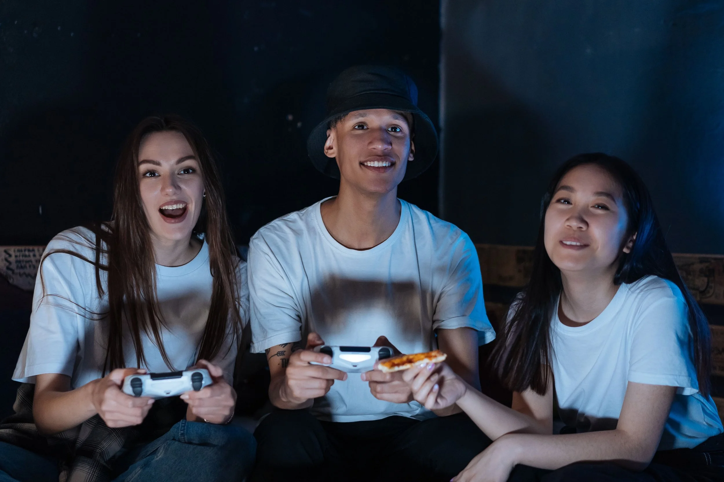 Four young friends sitting together in a dark room, playing video games and smiling.