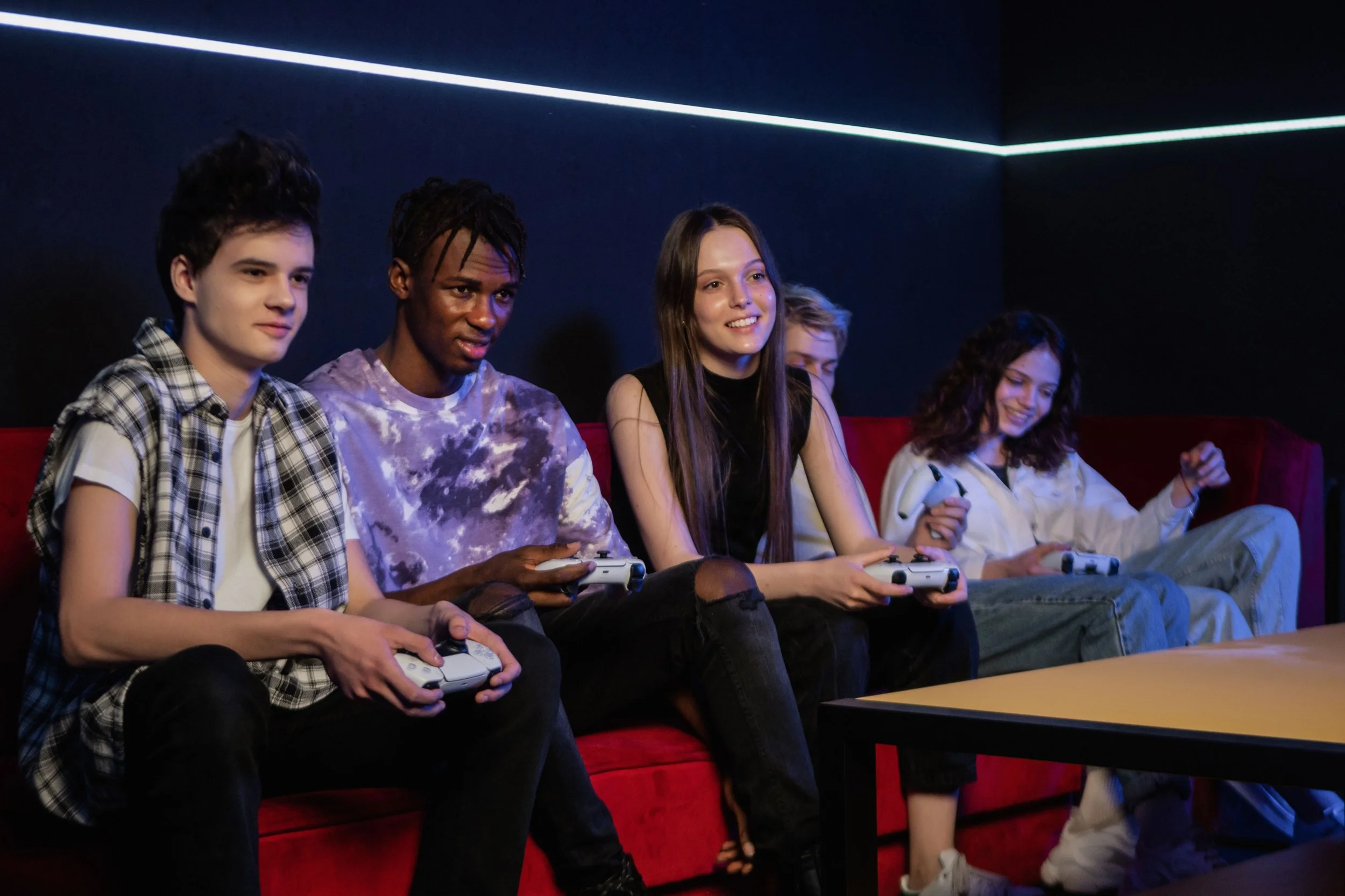 Group of five teenagers sitting on a red couch playing video games in a dark room with neon lighting.