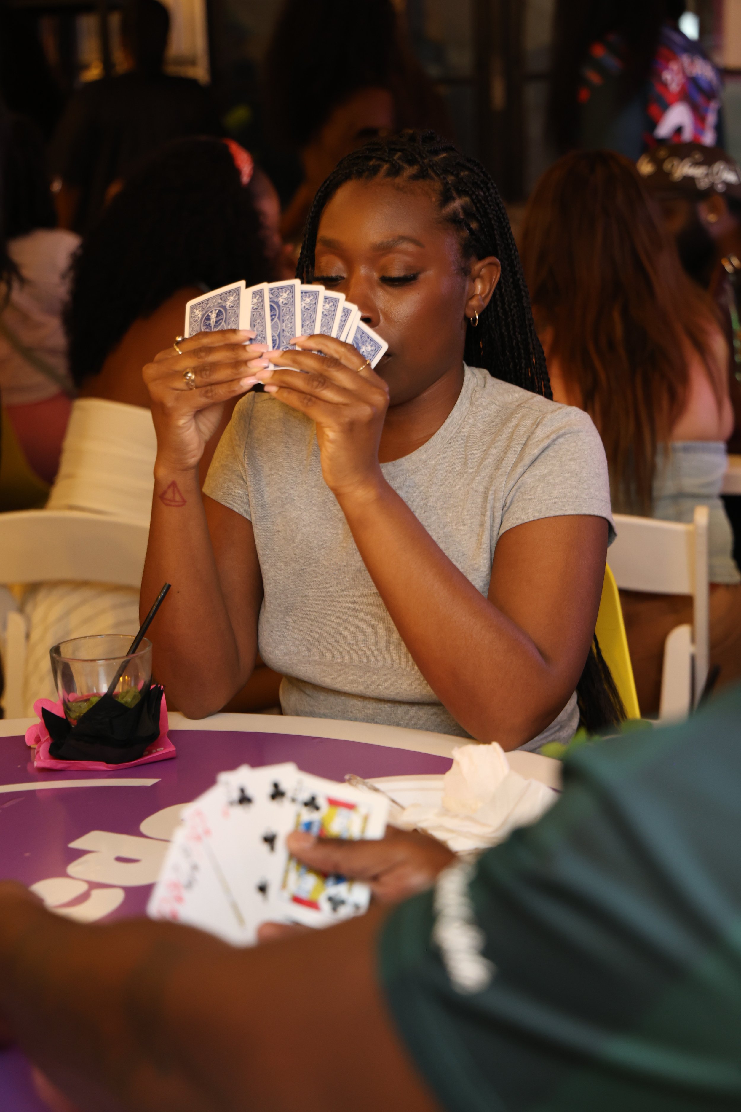 A woman with braided hair is holding and examining a hand of playing cards at a table during a gathering. Other people are visible in the background.