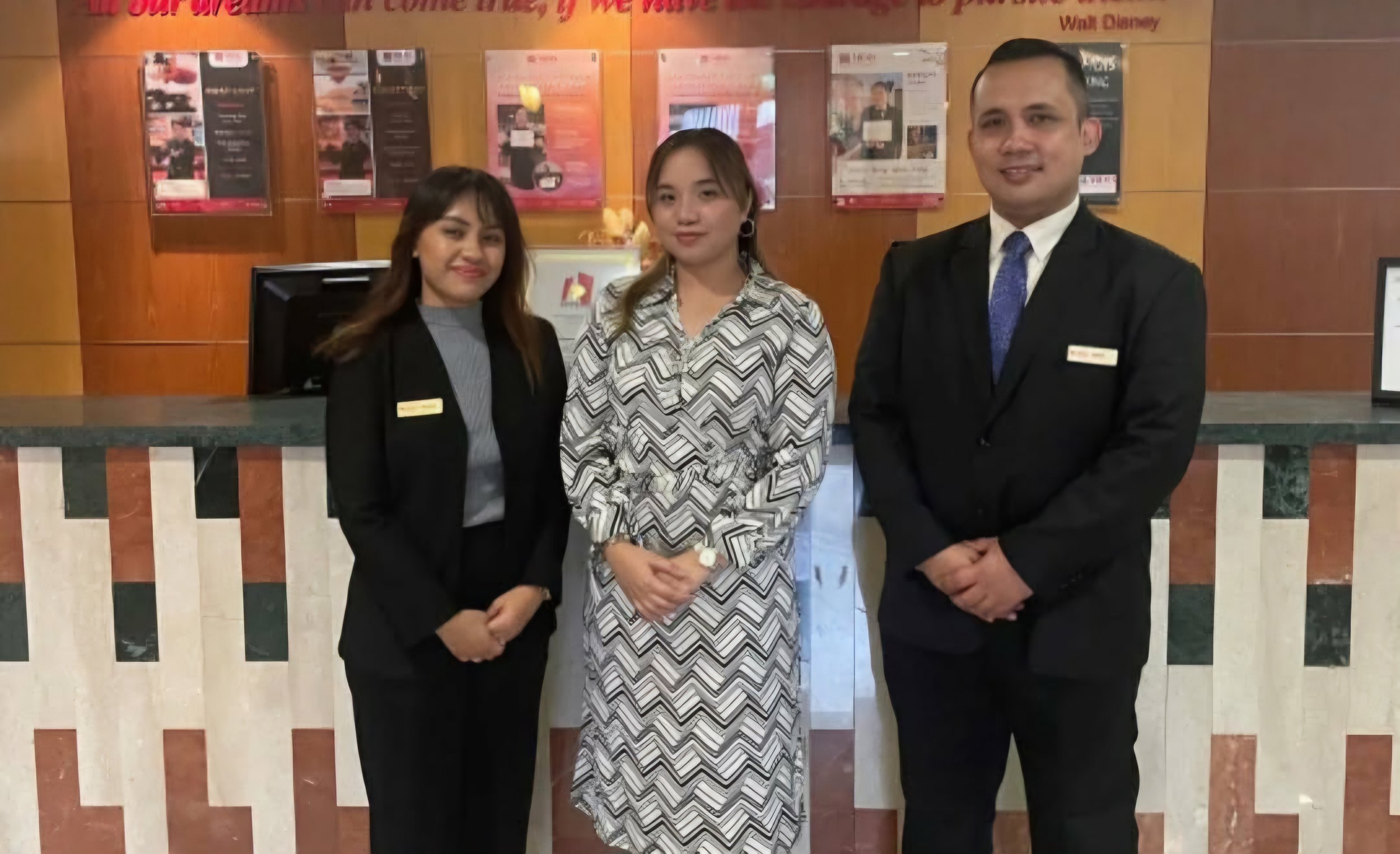 Three people standing behind a reception desk in a hotel lobby, smiling for a photo. The two women are dressed in formal attire and the man is wearing a suit with a blue tie.