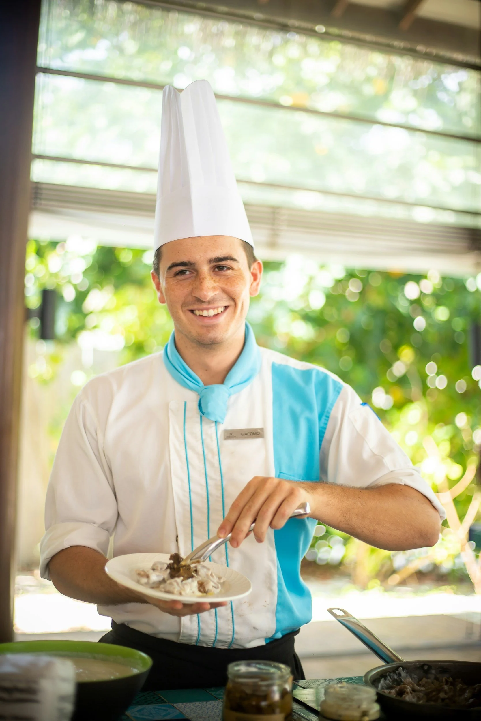 A smiling male chef wearing a traditional white chef hat and a blue and white uniform holds a plate of food while preparing a meal at an outdoor kitchen. There are bowls, jars, and utensils on the table.