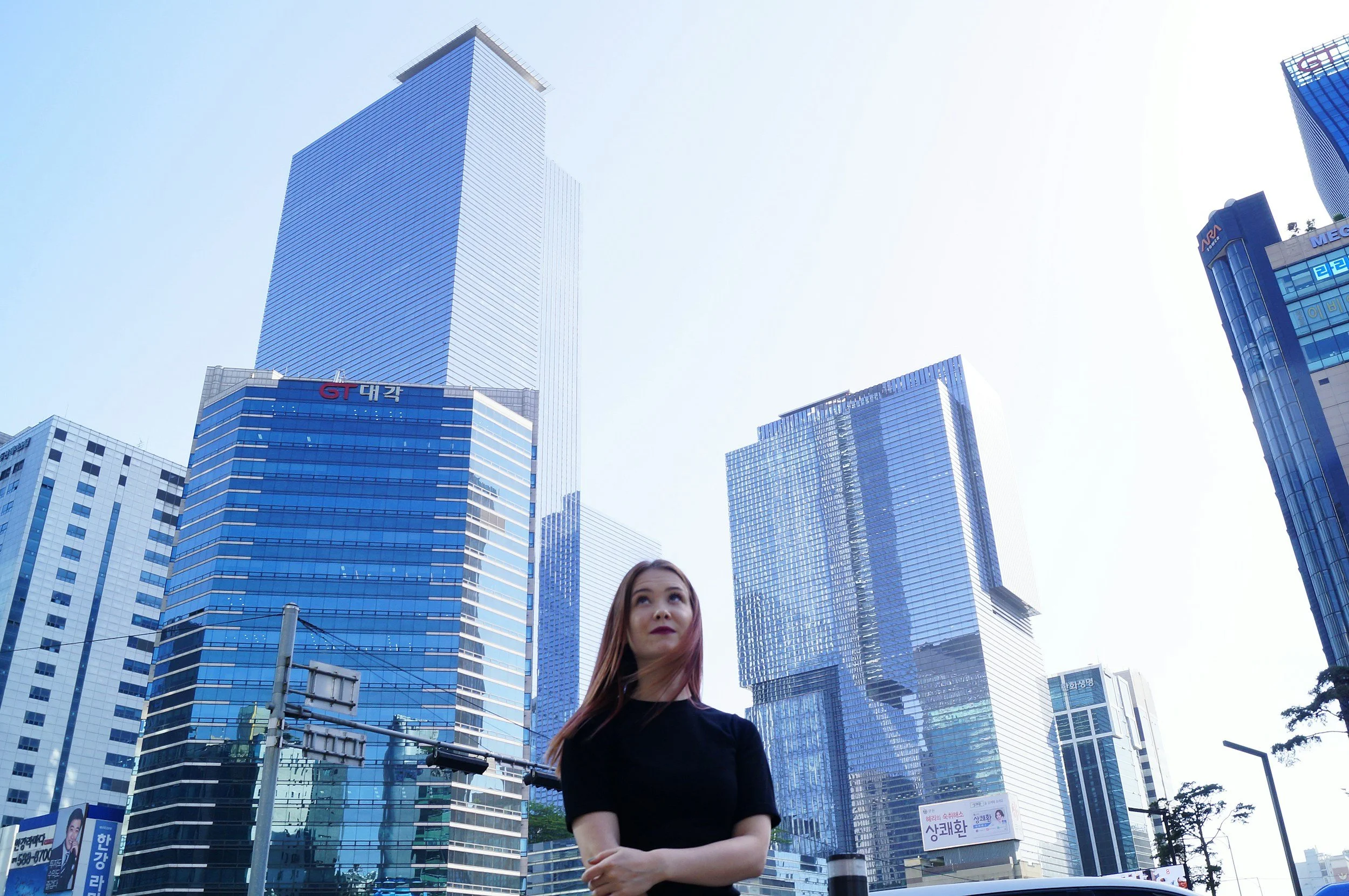 Woman with long red hair wearing a black top standing in front of tall modern glass skyscrapers in a city.