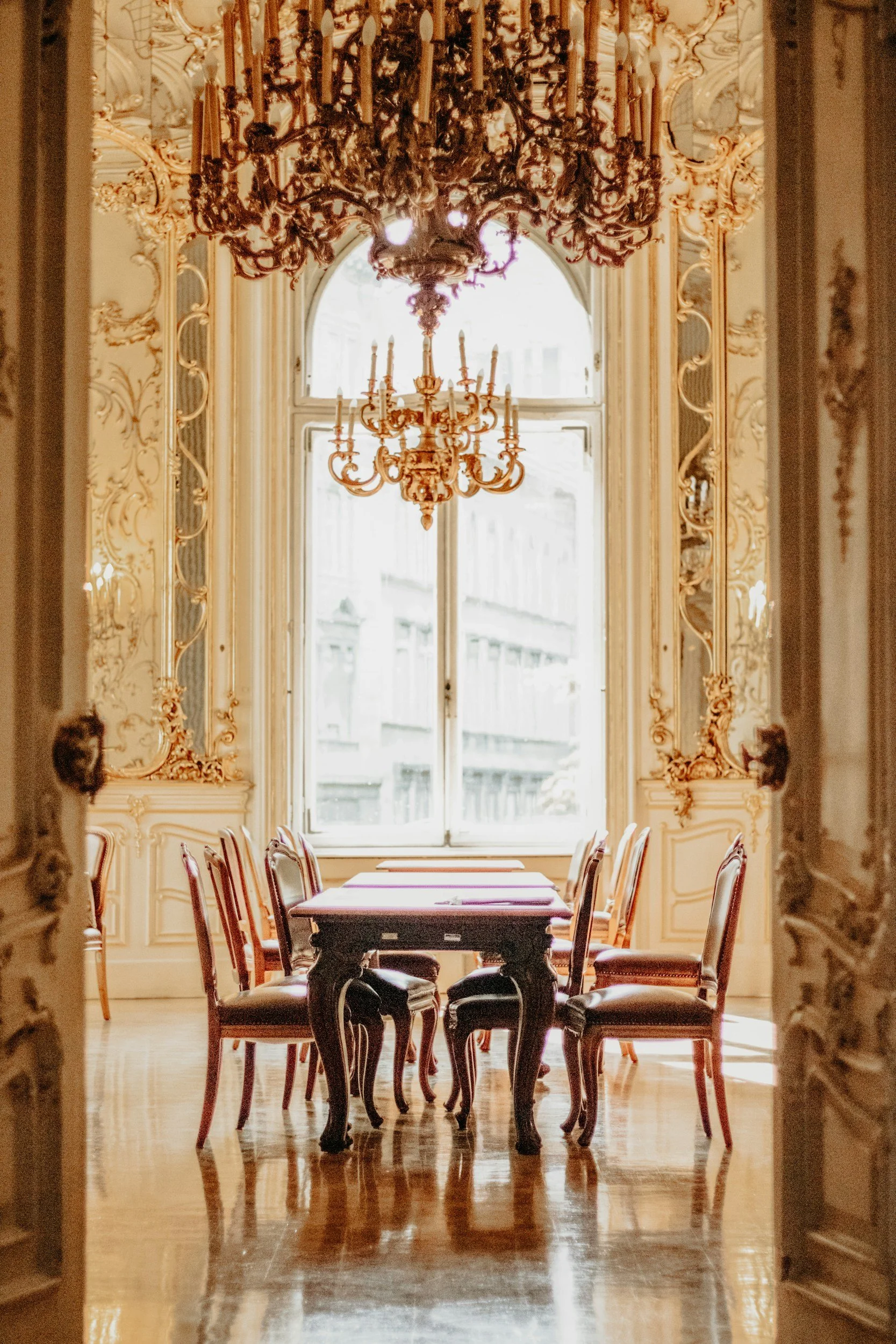 An ornate dining room with a long table and chairs, illuminated by a large chandelier and natural light from a big window with views of a cityscape.