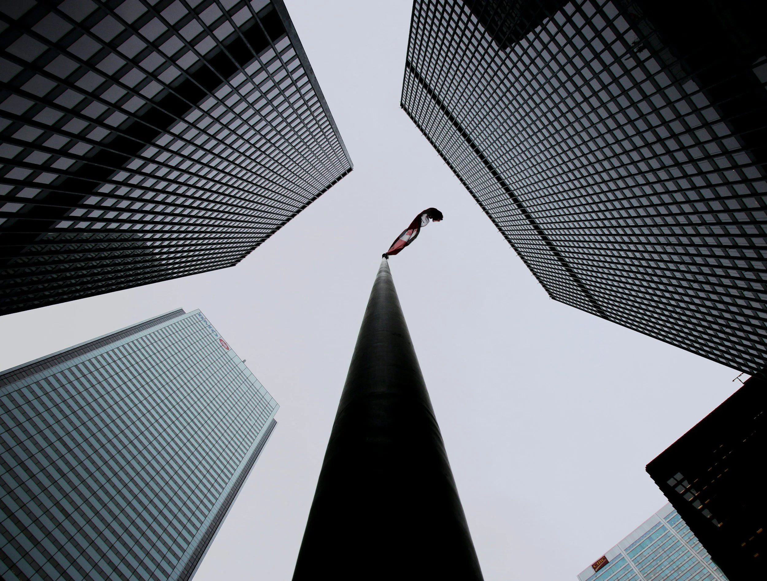 Looking up at tall skyscrapers with glass exteriors surrounding a flagpole with a flag in the middle.