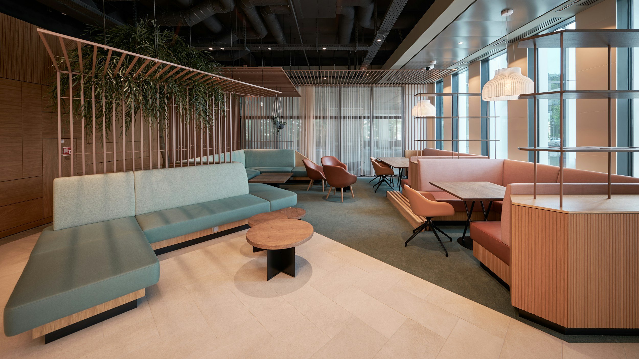 Modern indoor lounge area with pastel-colored seating, wooden furniture, large windows, and hanging pendant lights.