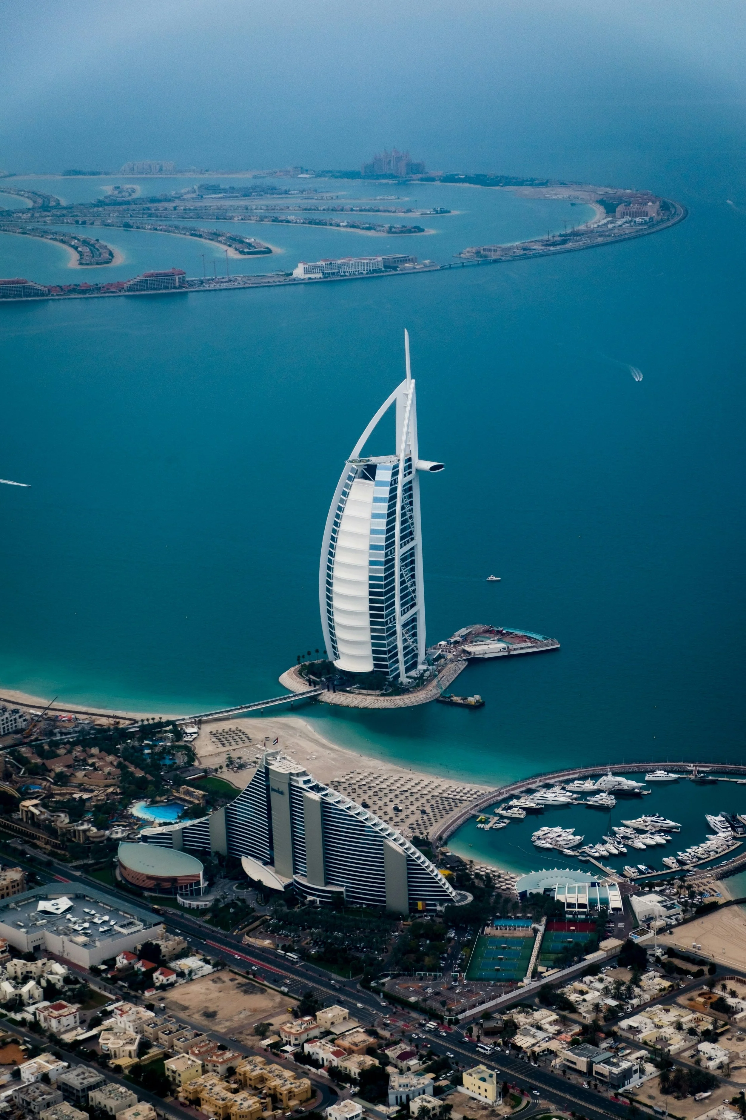 An aerial view of the iconic Burj Al Arab hotel in Dubai, shaped like a sailboat, surrounded by water and cityscape.