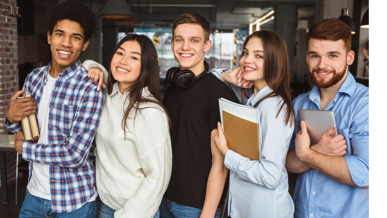 Group of five young adults smiling, standing close together in a modern, industrial-style office or cafe with brick walls and large windows in the background.