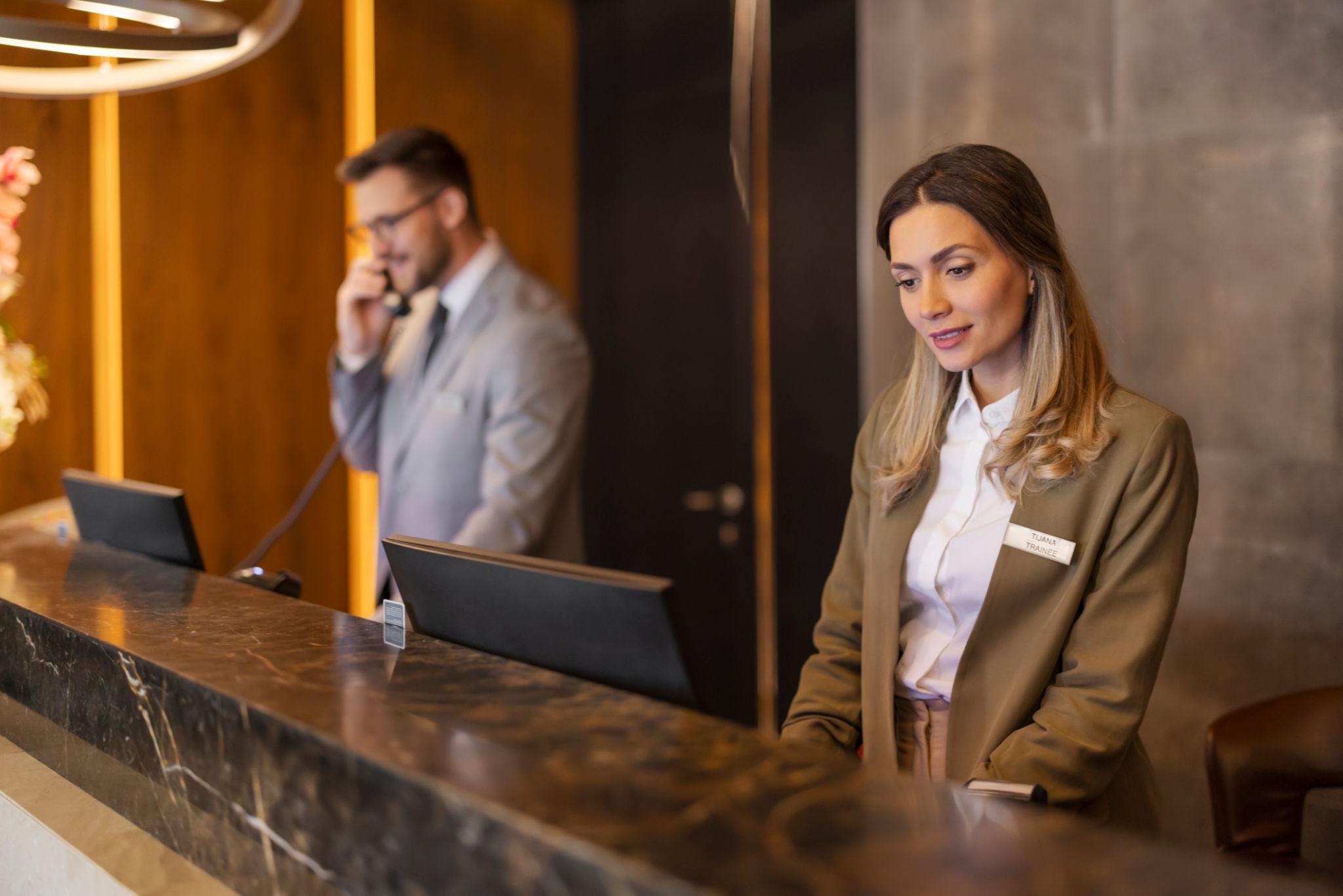 Hotel front desk with a woman receptionist wearing a beige blazer and white shirt, using a computer, and a man guest in a grey suit talking on a phone in the background.