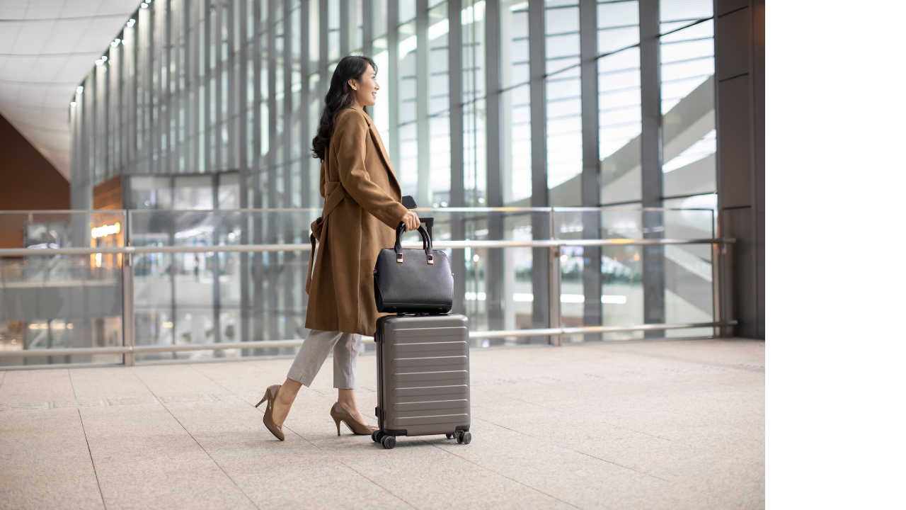 A woman in business attire with a coat and heels walking through an airport, pulling a rolling suitcase and carrying a handbag.
