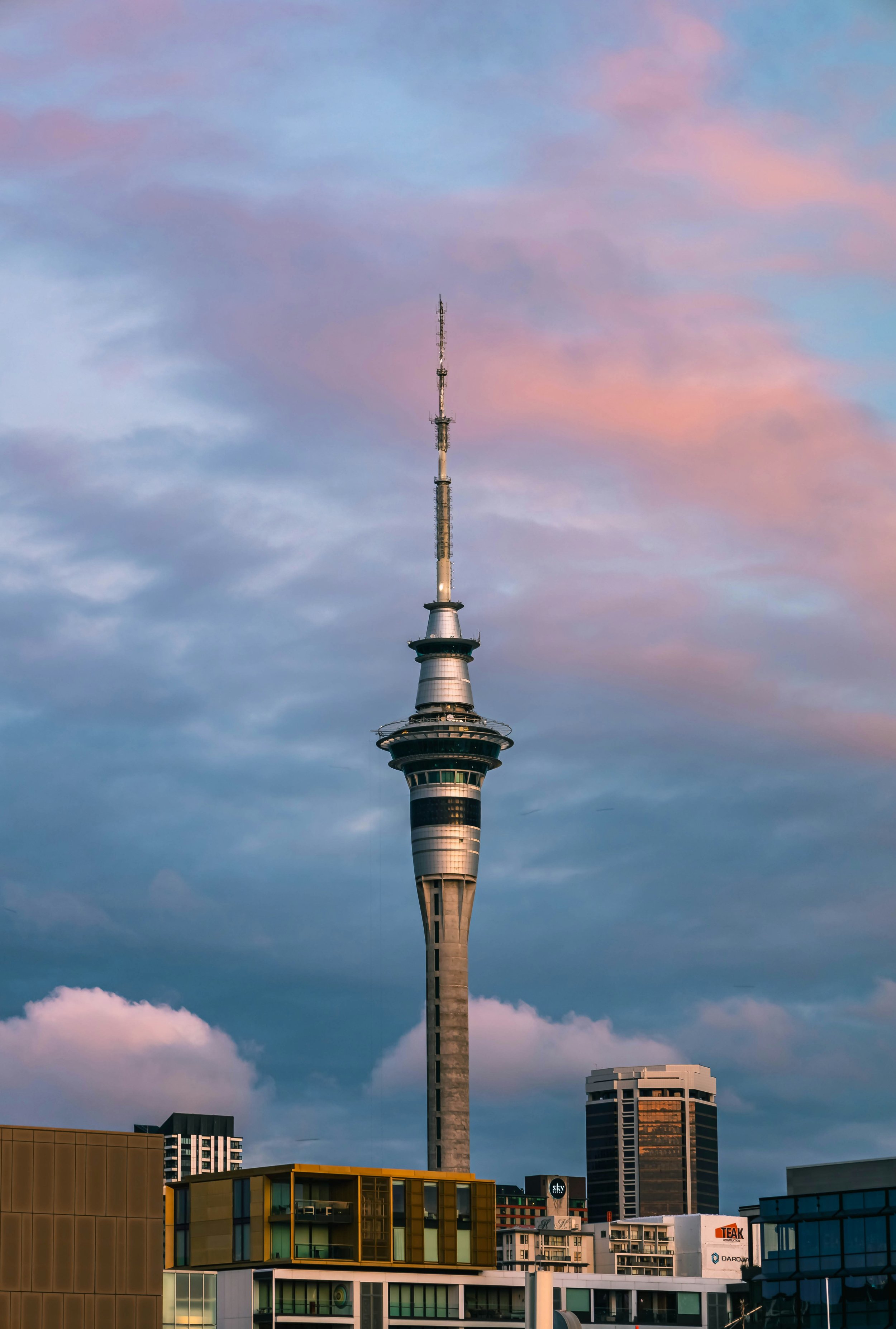 The Sky Tower, a tall telecommunications and observation tower, against colorful sunset sky with some clouds, surrounded by modern buildings in Auckland, New Zealand.