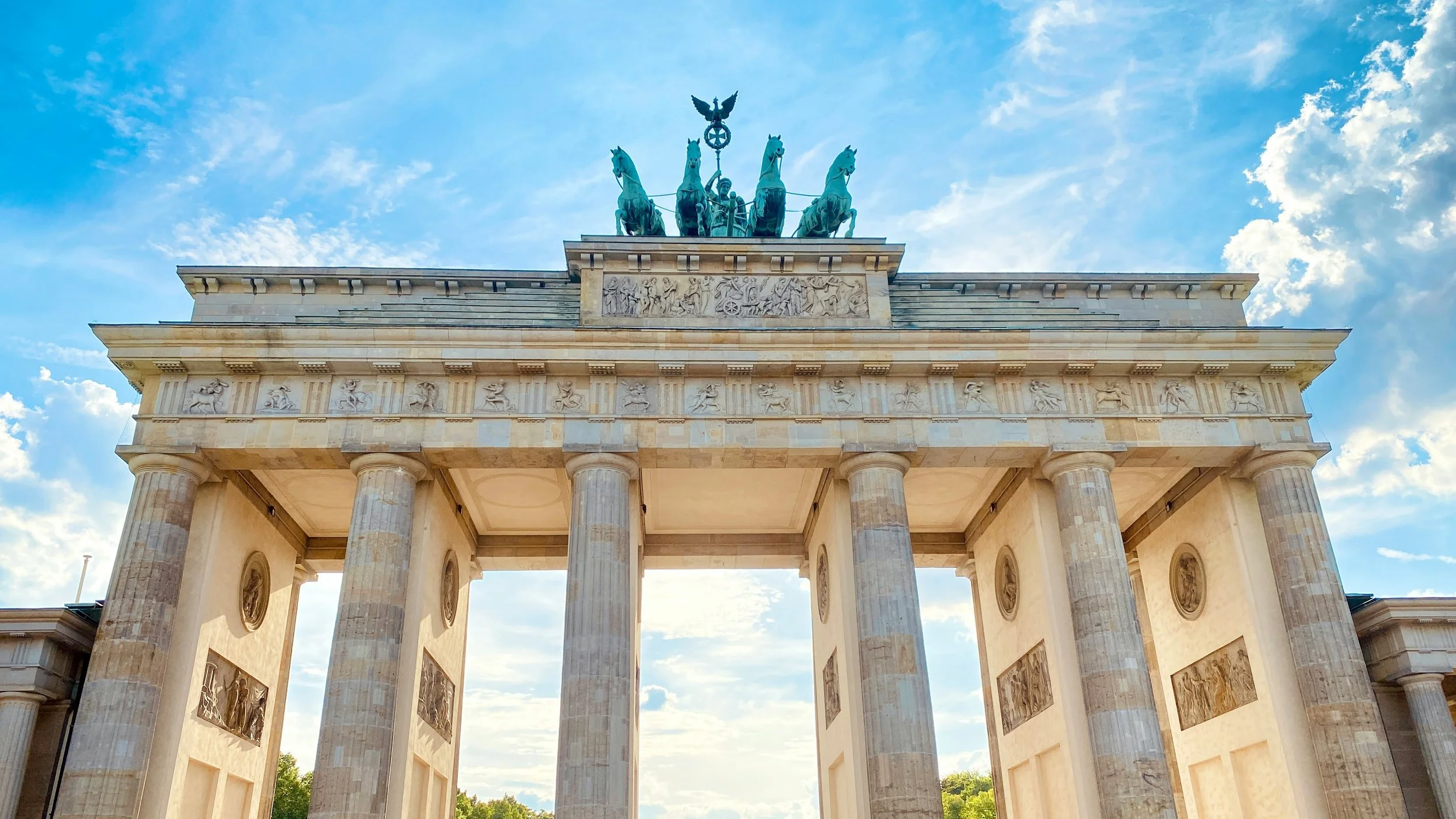 View of the Brandenburg Gate in Berlin, Germany, with blue sky and clouds above, showcasing neoclassical architecture and a quadriga statue at the top.
