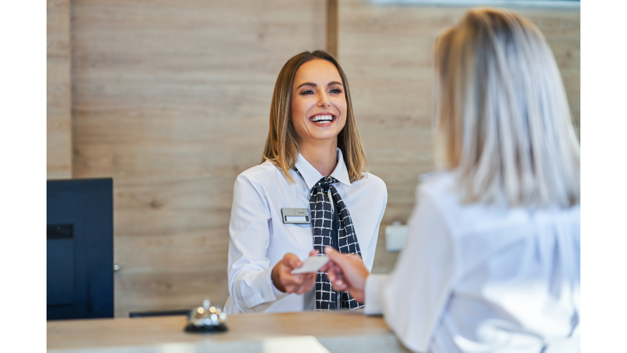 Receptionist smiling and handing a card to a guest at a hotel desk, with a wooden wall in the background.