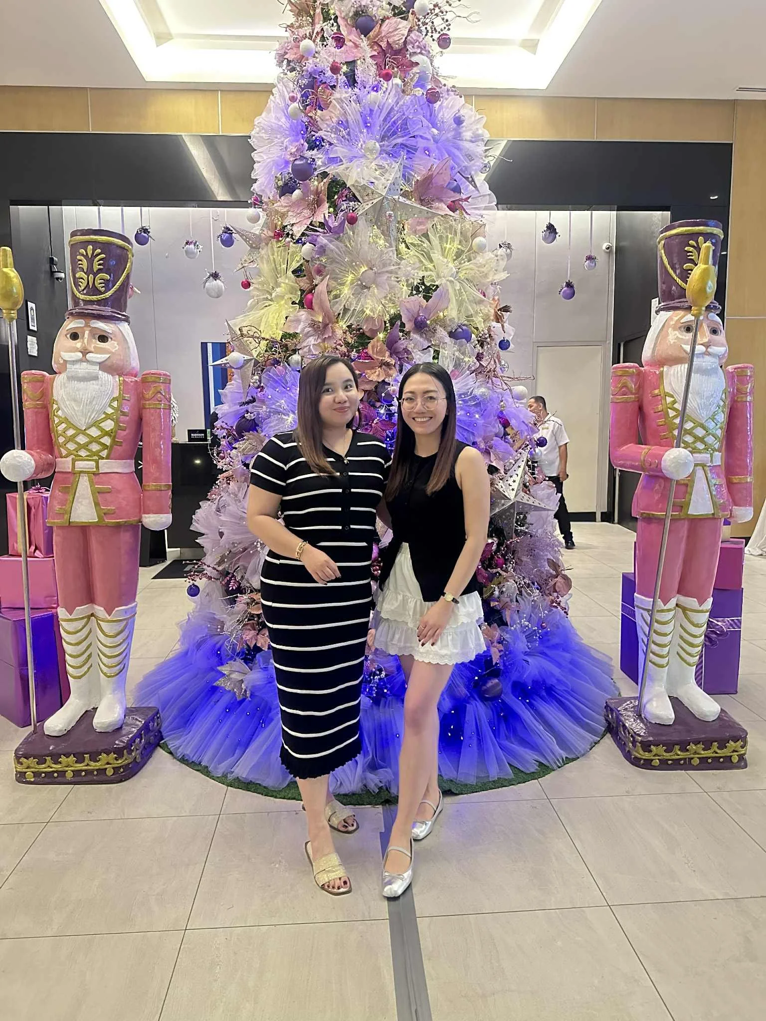 Two women stand in front of a decorative Christmas display with large pink and purple nutcracker statues on either side and a tall, colorful Christmas tree behind them. The women are smiling and wearing fashionable clothing, one in a striped dress and the other in a black top and white skirt.