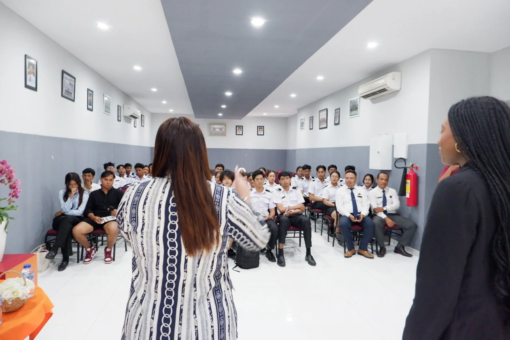 A group of students in white uniforms attending a presentation in a classroom or meeting room, with two women standing at the front speaking to the audience.