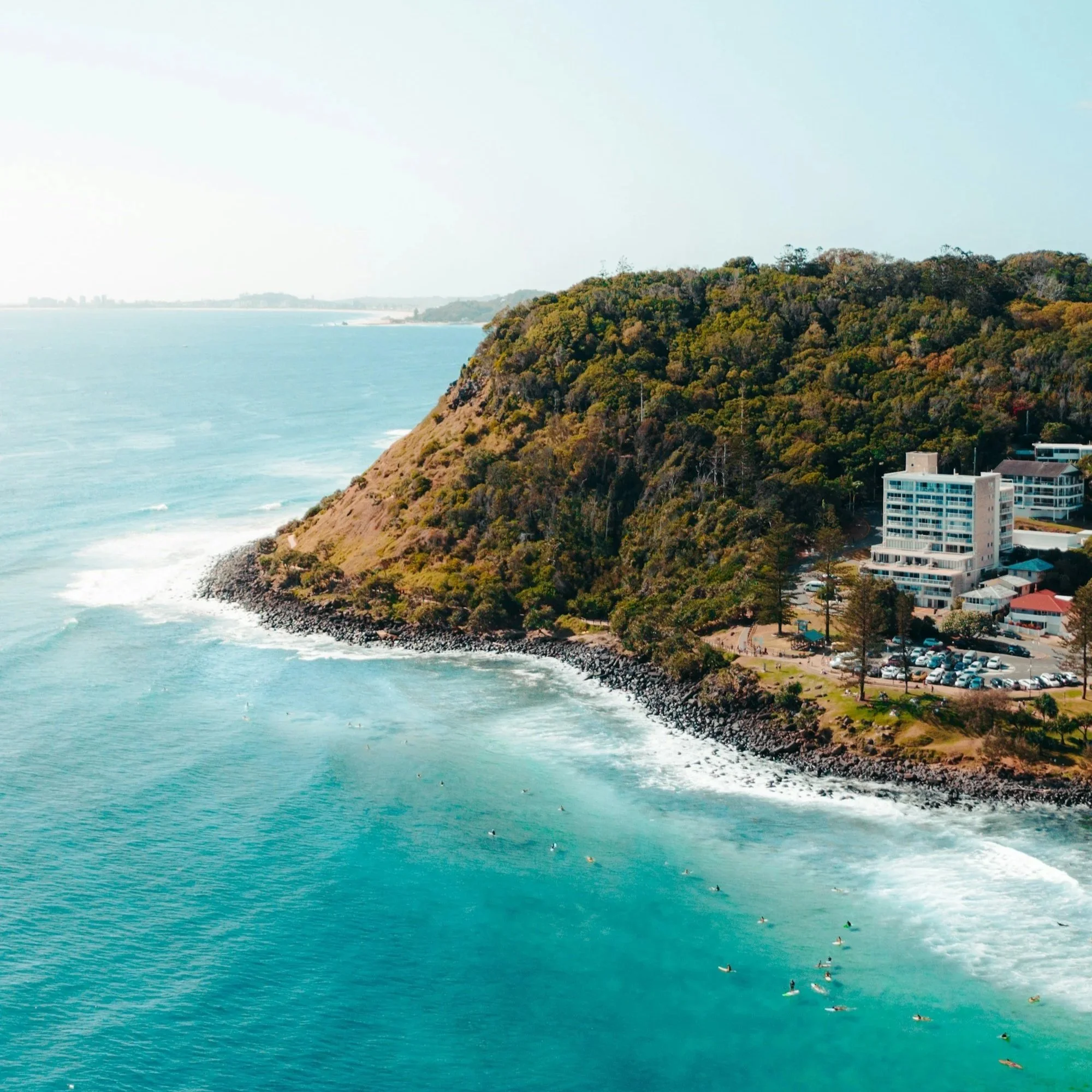 A coastal landscape featuring a hillside covered with trees, overlooking the ocean with waves and a rocky shoreline, with residential buildings at the water's edge.