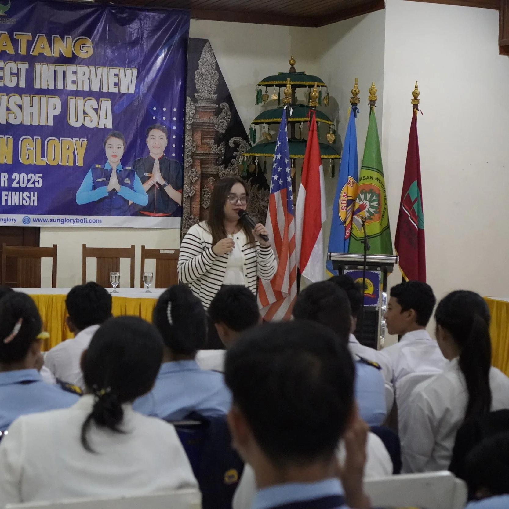 A woman speaker holding a microphone presents in front of a group of students in uniforms, seats facing her. Behind, a banner with text and images, and several flags are displayed.