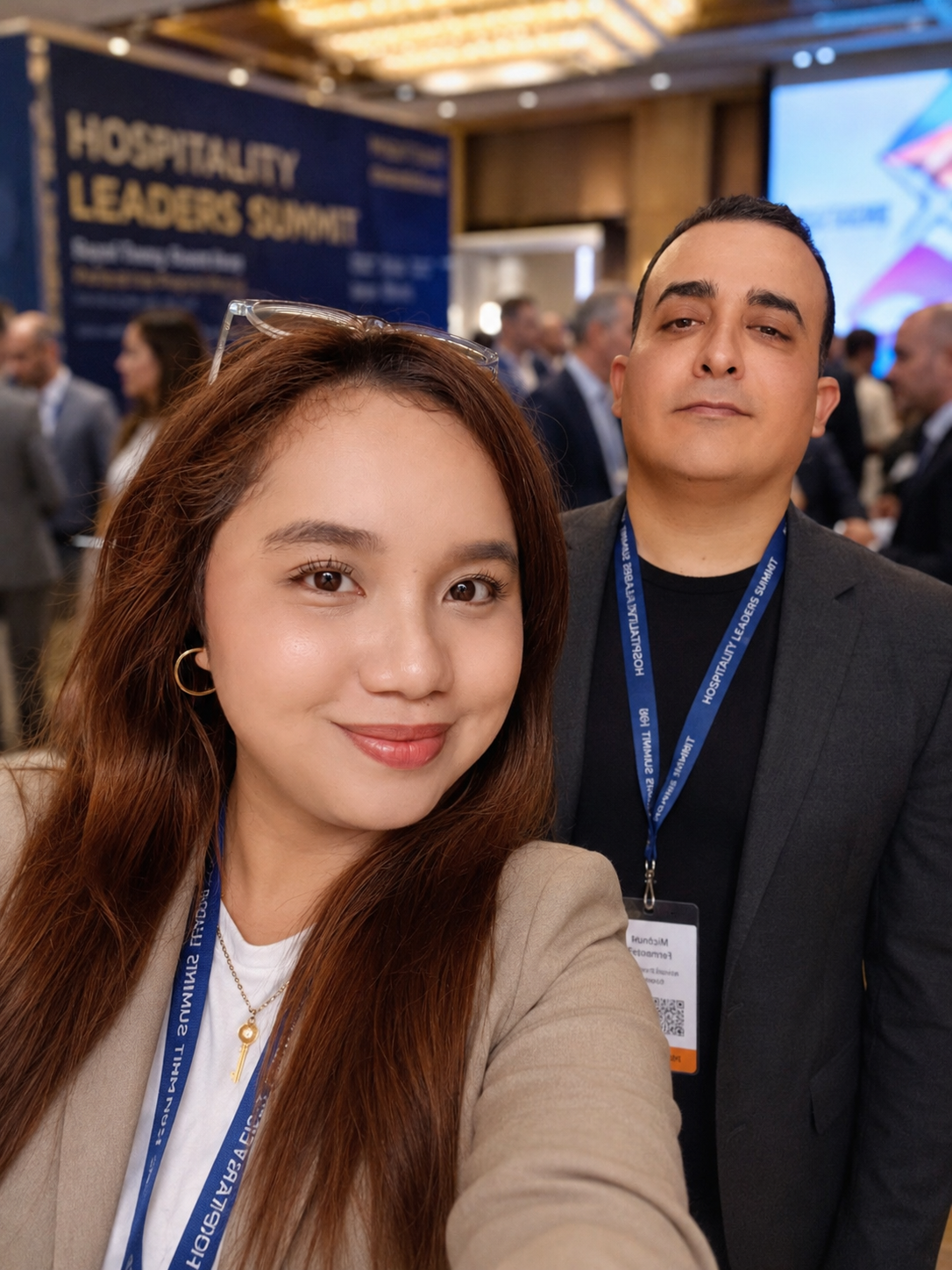 Selfie of a woman with long reddish-brown hair and hoop earrings smiling, and a man with short dark hair and a neutral expression, both wearing conference badges and lanyards at the Hospitality Leaders Summit event.