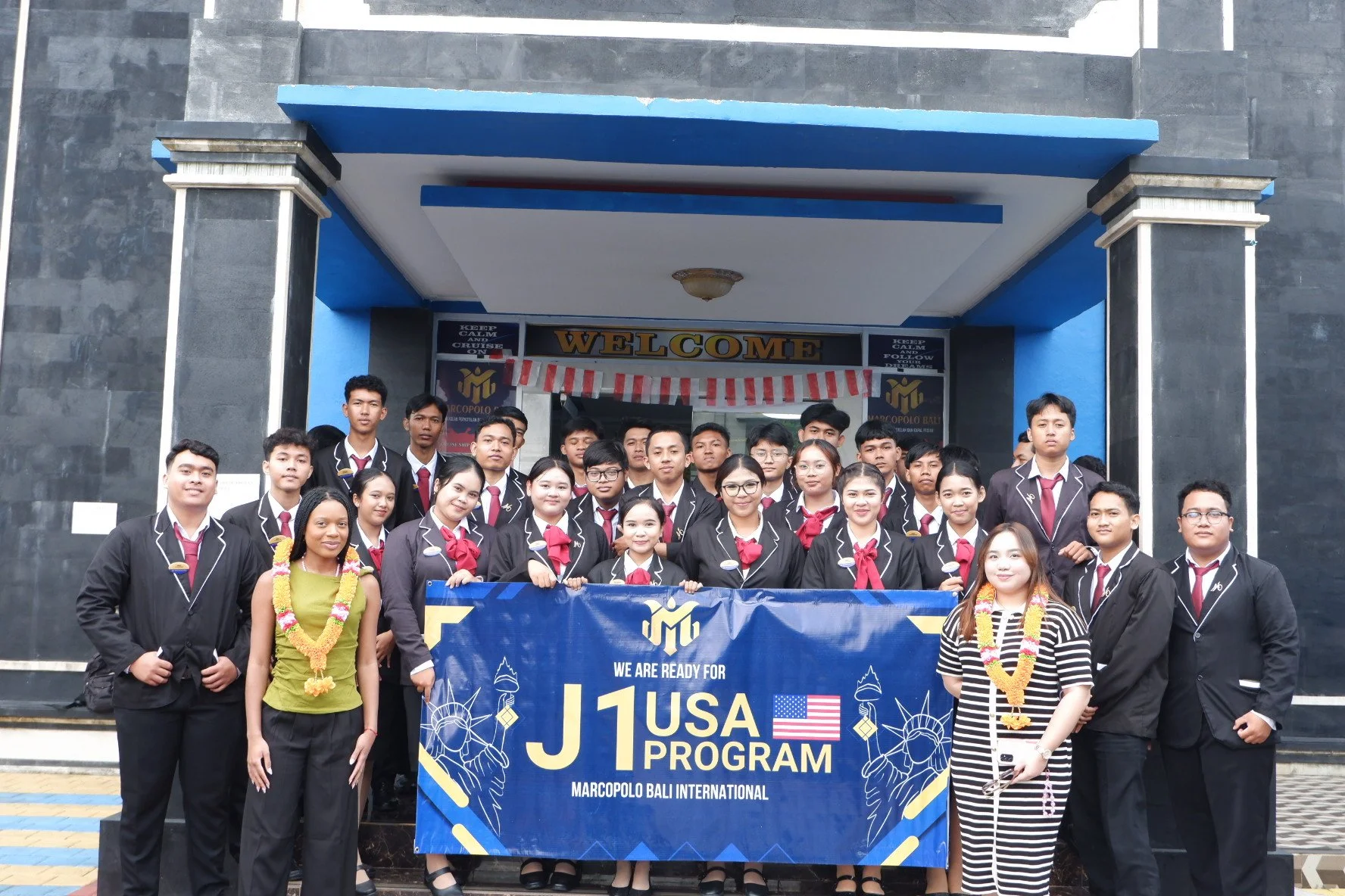 Group of young students in black uniforms with red ties and white shirt collars, standing in front of a building, holding a blue banner that reads "J1 USA Program" and "Marcopolo Bali International," with some wearing traditional flower garlands, posing for a photo during a student exchange program.