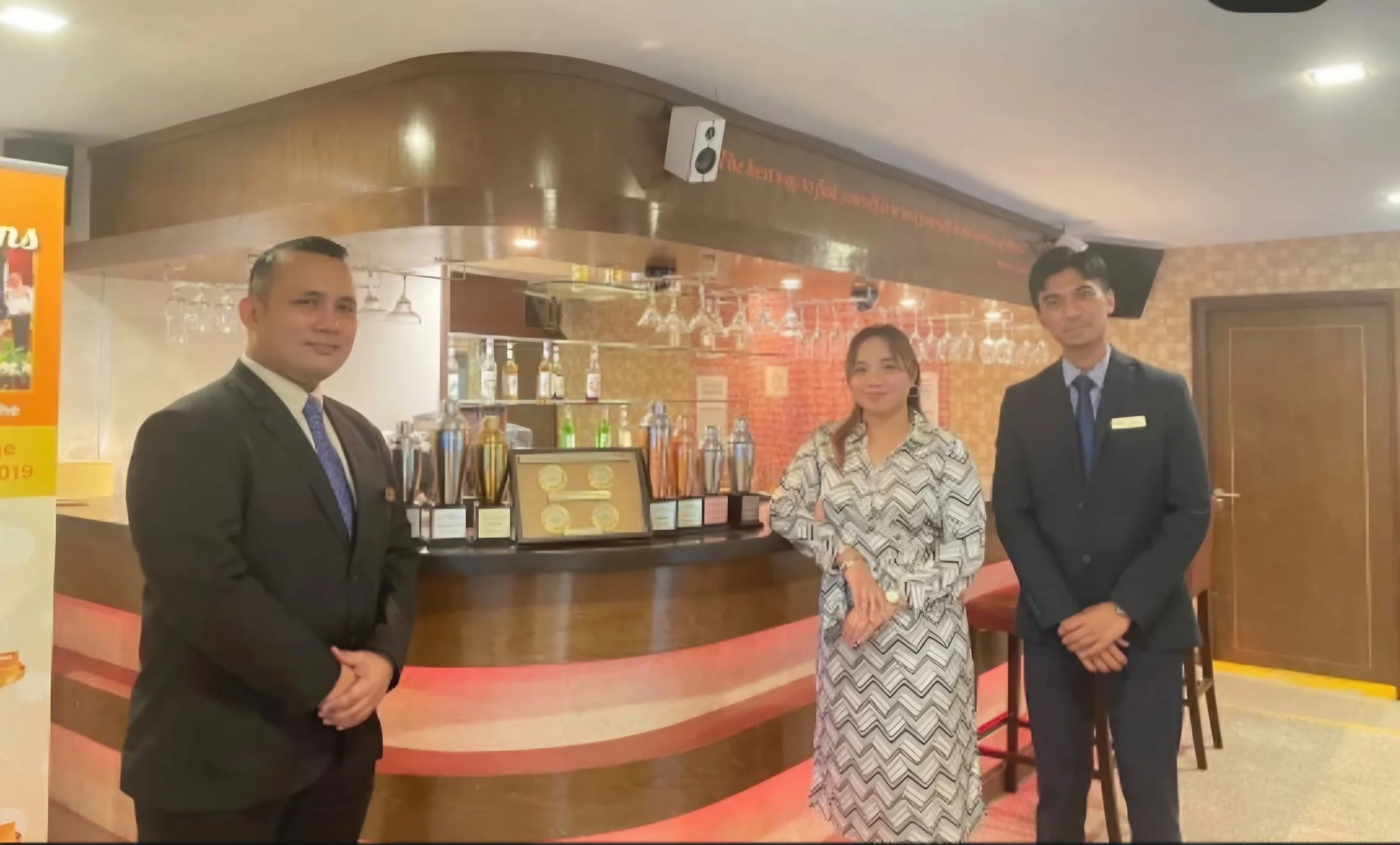 People standing in front of a reception desk in a restaurant or hotel lobby, with some awards or plaques displayed on the counter.