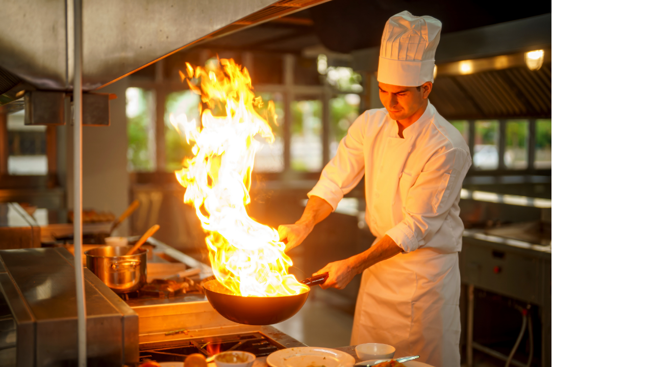 Chef in white uniform and hat cooking flambé in a professional kitchen with flames rising from a pan.