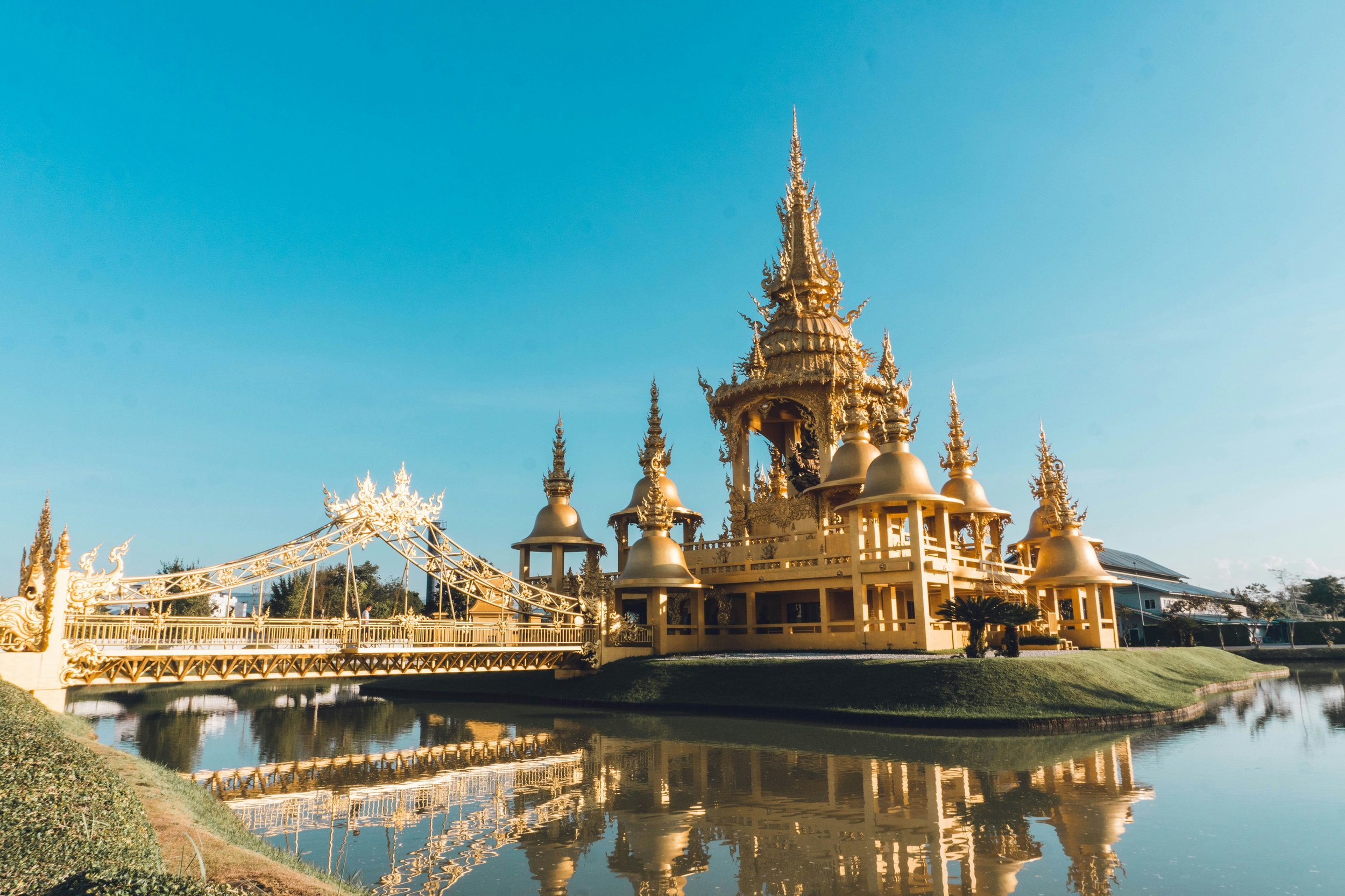 A golden ornate temple with a tall spire, surrounded by smaller structures, reflected in a calm body of water, under a clear blue sky.