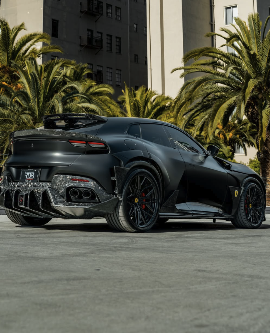 A matte black Ferrari sports car parked on the street with palm trees and a building in the background.