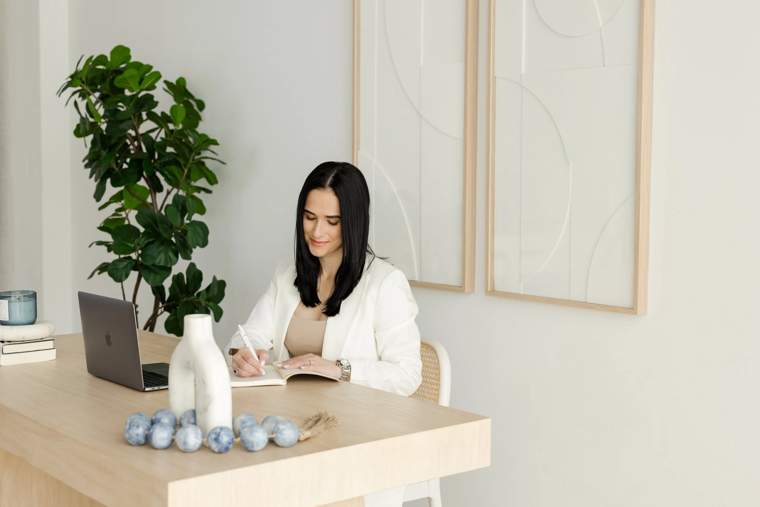 A woman with black hair and a white blazer sitting at a wooden table in a bright, modern room, writing in a notebook while working on a laptop.