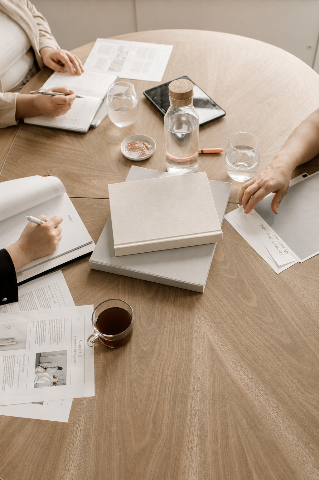 People sitting around a wooden table with notebooks, papers, a tablet, glasses of water, a bottle of water, a cup of tea, and a few closed boxes.