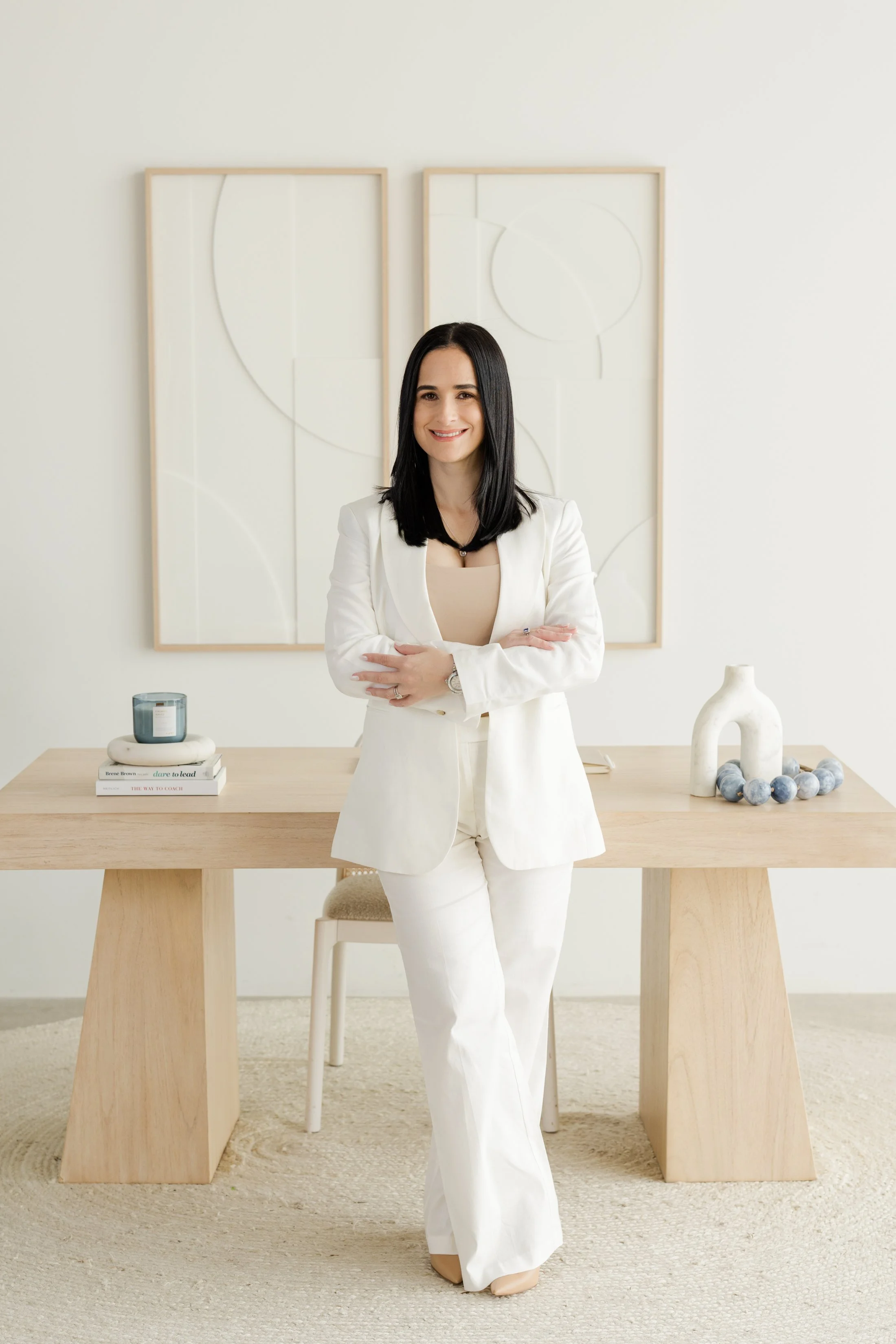 A woman with black hair, wearing a white blazer and matching pants, standing with arms crossed in a minimalist room with a light wood table and neutral decor.