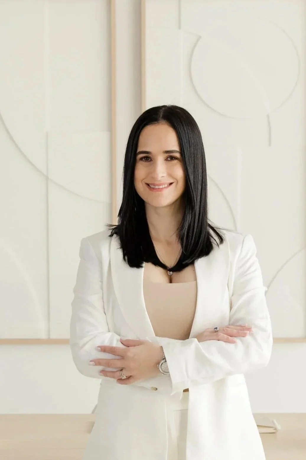 A woman with black hair and light skin, smiling, wearing a white blazer and beige top, standing with arms crossed in an office or modern room with white walls and decorative circular artwork.