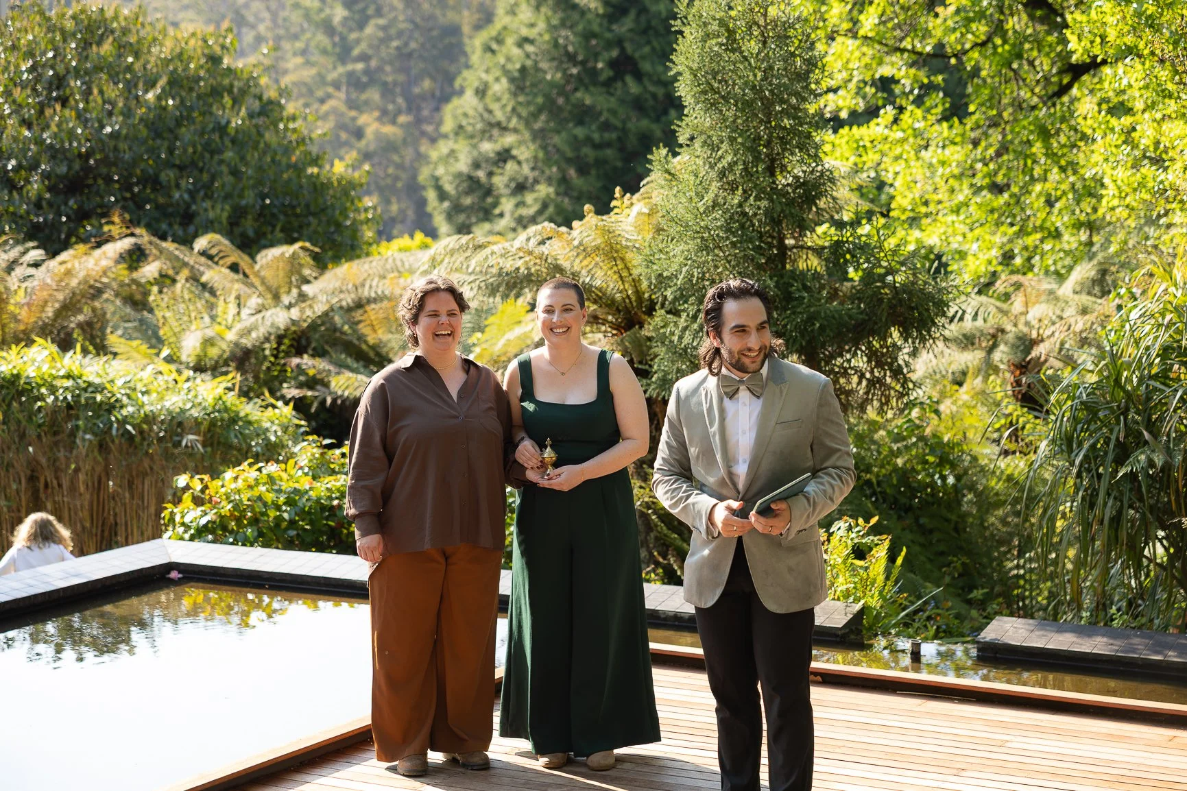 Three people, two women and one man, standing outdoors near a pond and lush green trees, smiling and engaged in conversation on a sunny day.