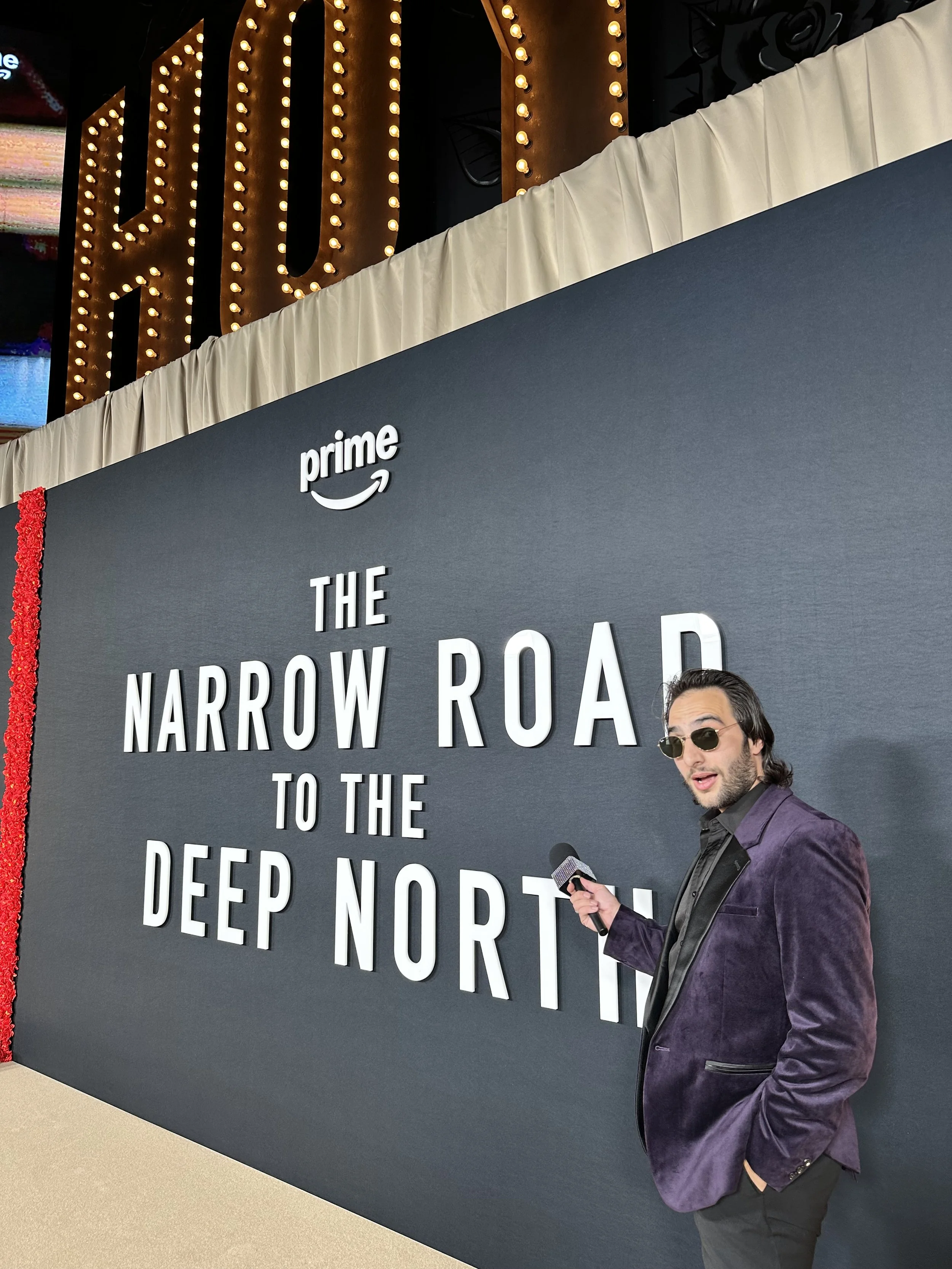A man in a purple velvet blazer and black shirt, wearing sunglasses, stands in front of a large black sign with white text and the Amazon Prime logo, at what appears to be an event or press conference.