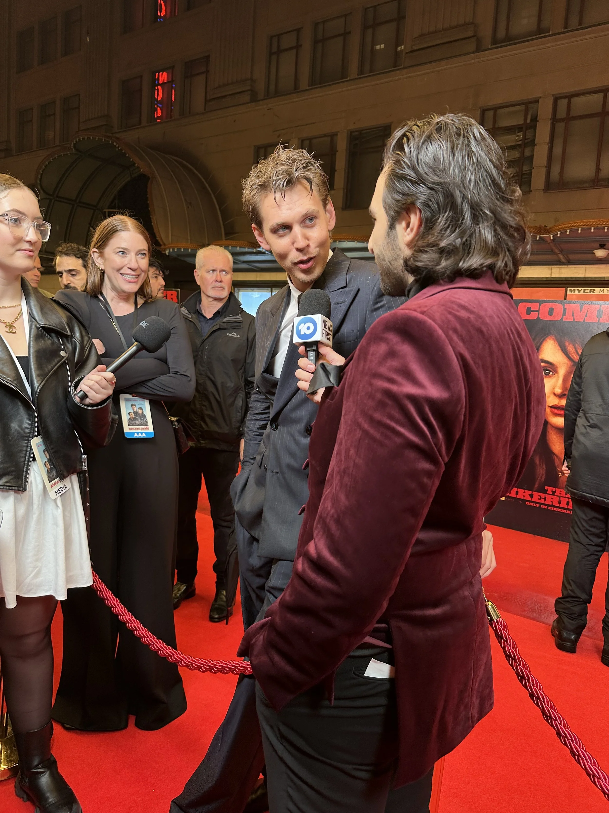 Two men having an animated conversation at a red carpet event, surrounded by several women and men, with a poster of a movie in the background.