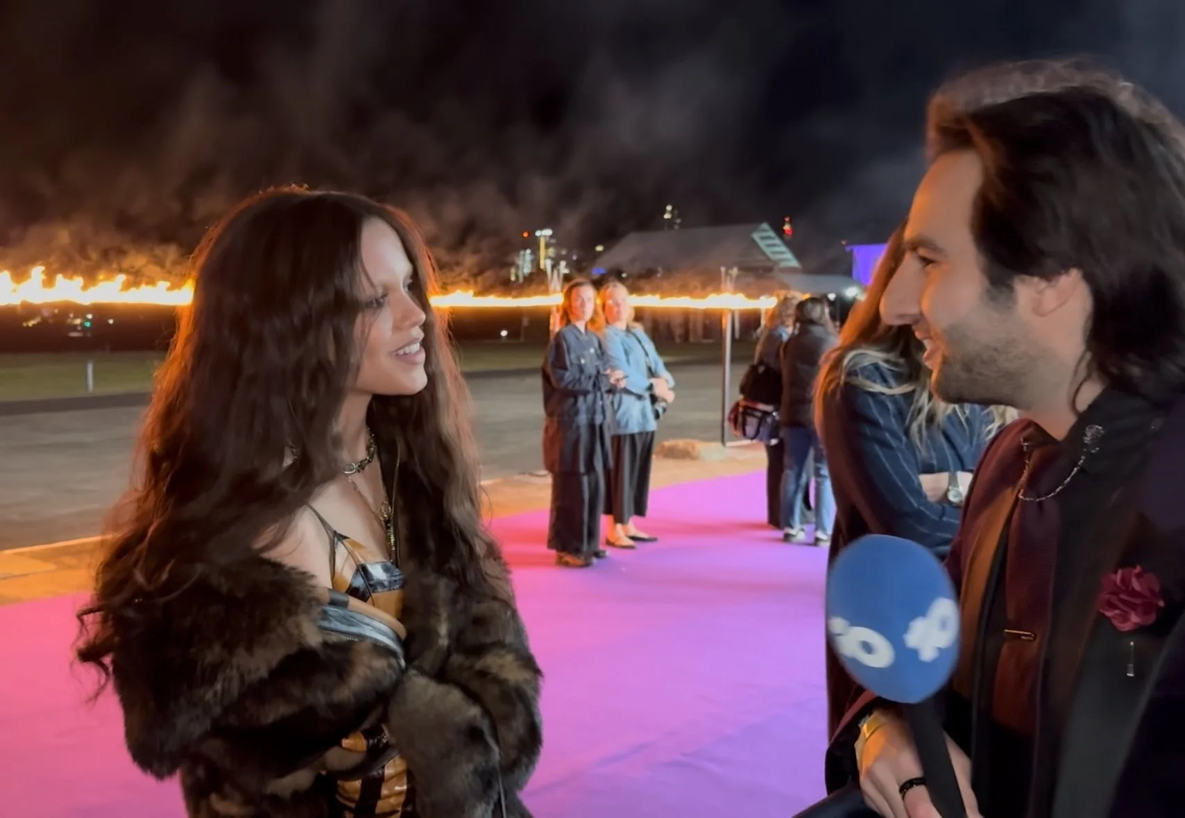 A woman and a man are facing each other and smiling at a nighttime event outdoors with smoke and fire in the background. There are a few women standing behind them on a pink carpet, and a city skyline can be seen in the distance.