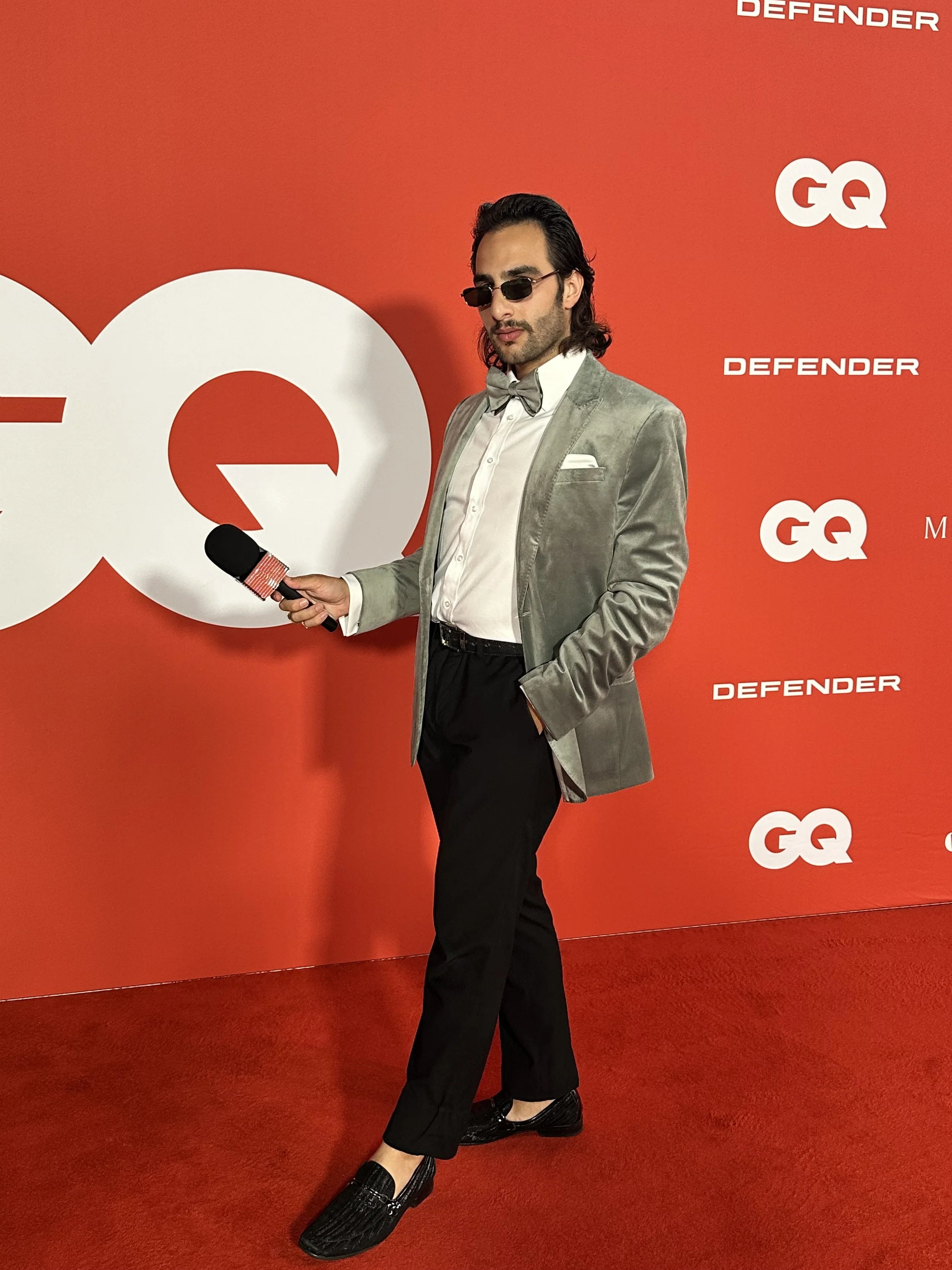 A man with dark hair, wearing sunglasses, a light gray velvet blazer, white shirt, gray bow tie, black pants, and black loafers, stands on a red carpet with a red backdrop featuring white logos of GQ and Defender, holding a microphone.