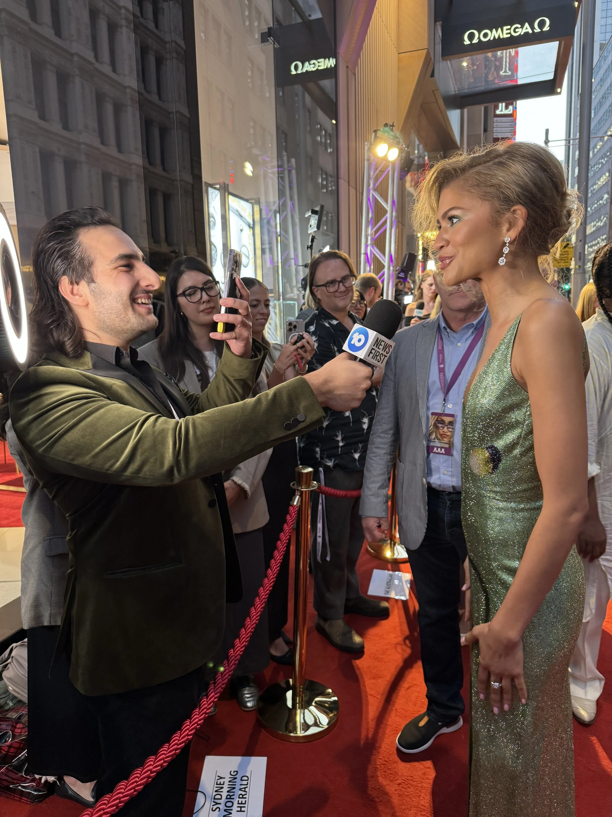 A woman in a sparkling green dress being interviewed by a man holding a microphone with the logo '10 NEWS FIRST' at a red carpet event outside a building in New York City.