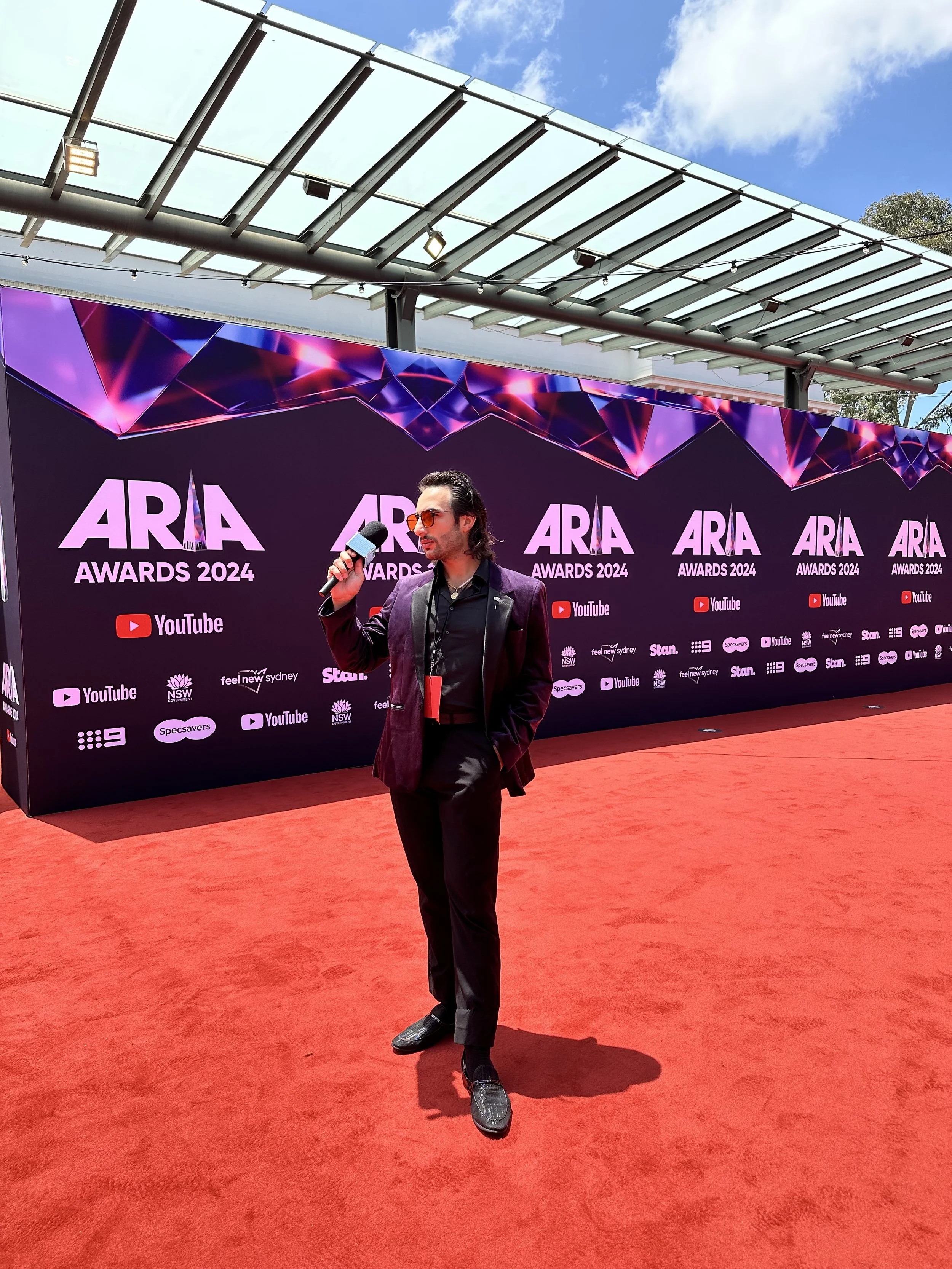 A man wearing dark sunglasses, a dark purple blazer, and black pants stands on a red carpet holding a microphone at the ARIA Awards 2024. Behind him, there is a large black background with pink and purple geometric patterns and logos including YouTube and other sponsors.