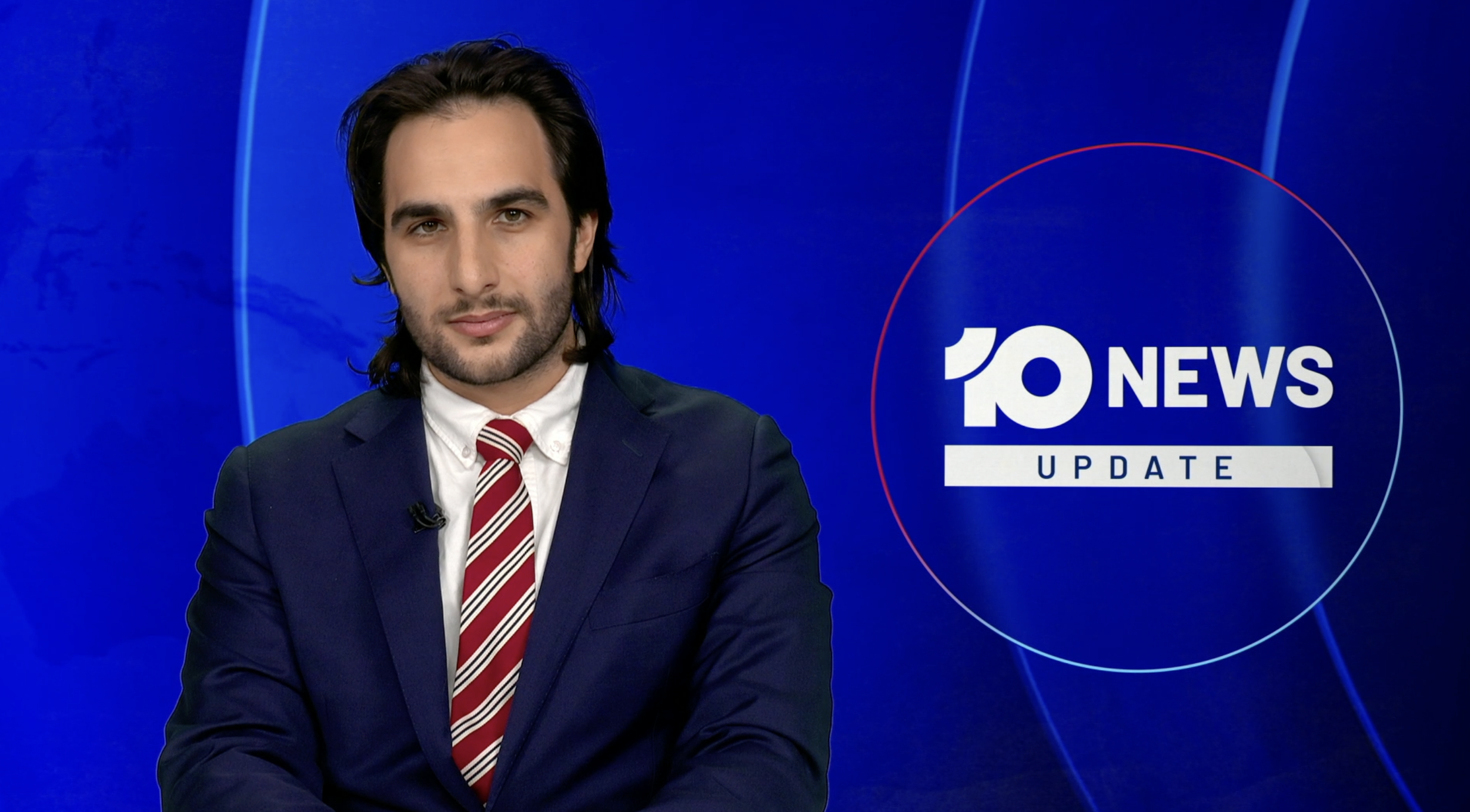 A news anchor with long dark hair wearing a navy suit, white shirt, and red striped tie, sitting at a news desk with a blue background displaying the '10 News Update' logo.