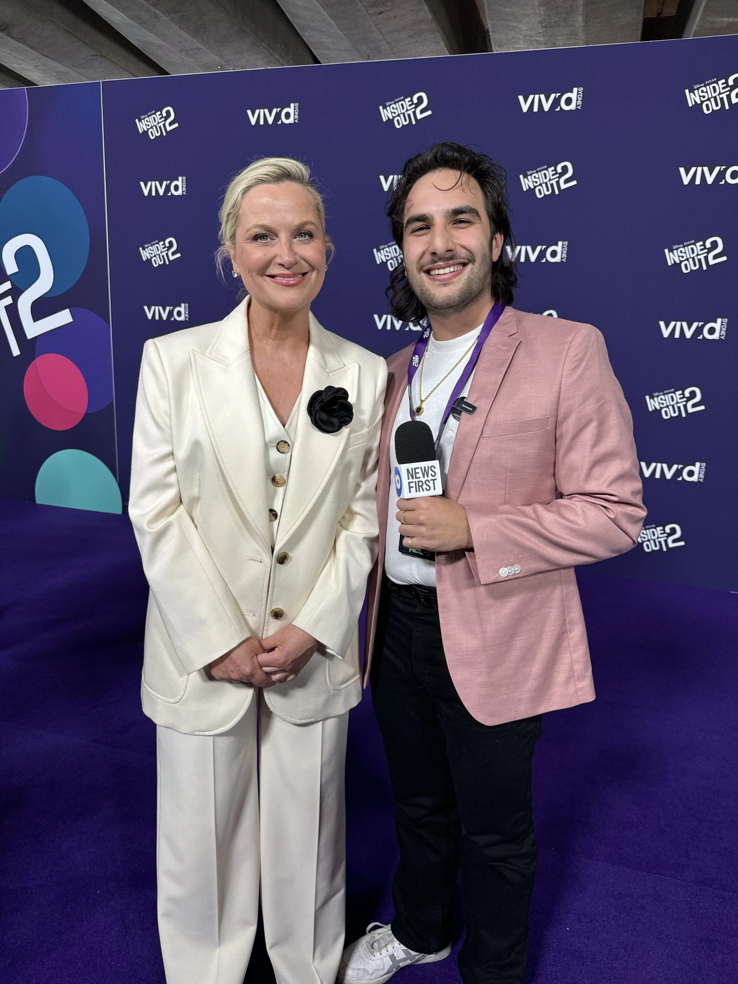 A woman in a cream-colored suit with a black flower pin stands next to a man in a pink blazer with a news microphone, in front of a backdrop with "Inside Out 2" and "VIV'ld" logos.