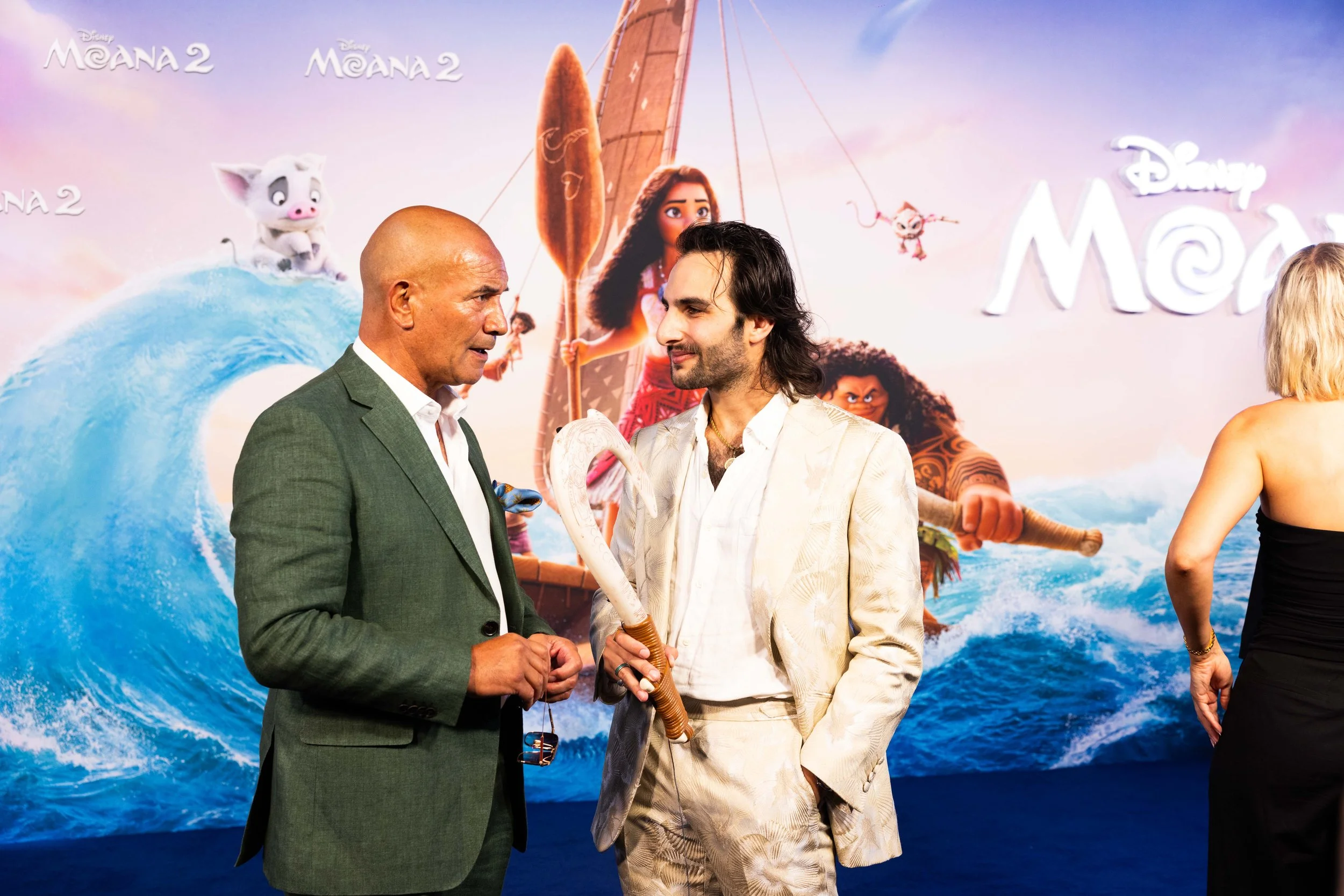 Two men standing in front of a colorful poster for Disney's Moana 2, having a conversation. The man on the left is bald and dressed in a green suit, while the man on the right has long dark hair and is dressed in a light-colored suit. The poster feat
