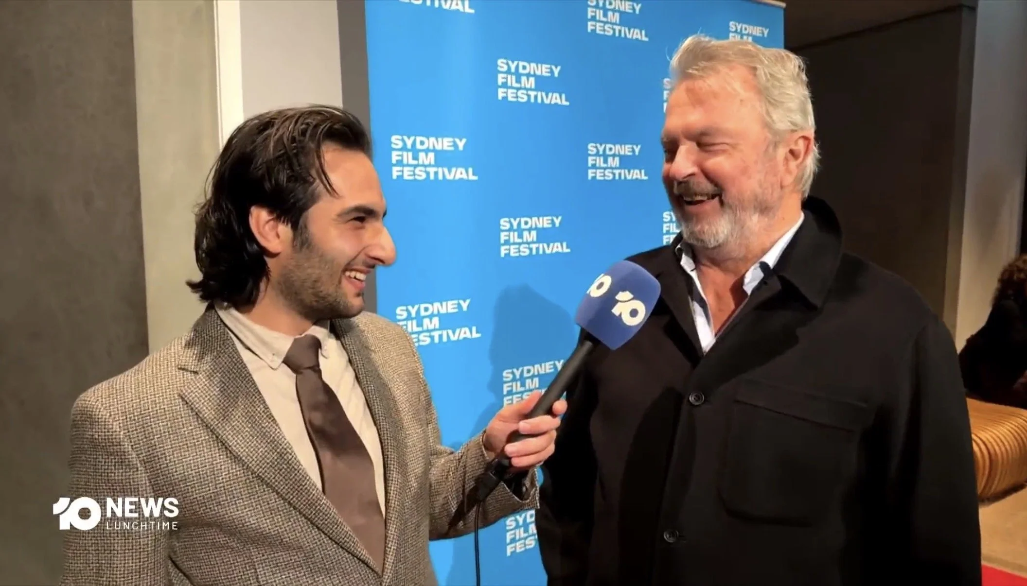 Two men smiling and talking during an interview at the Sydney Film Festival, with a blue backdrop displaying the festival name. One man is holding a microphone labeled '10 NEWS LUNCHTIME.'
