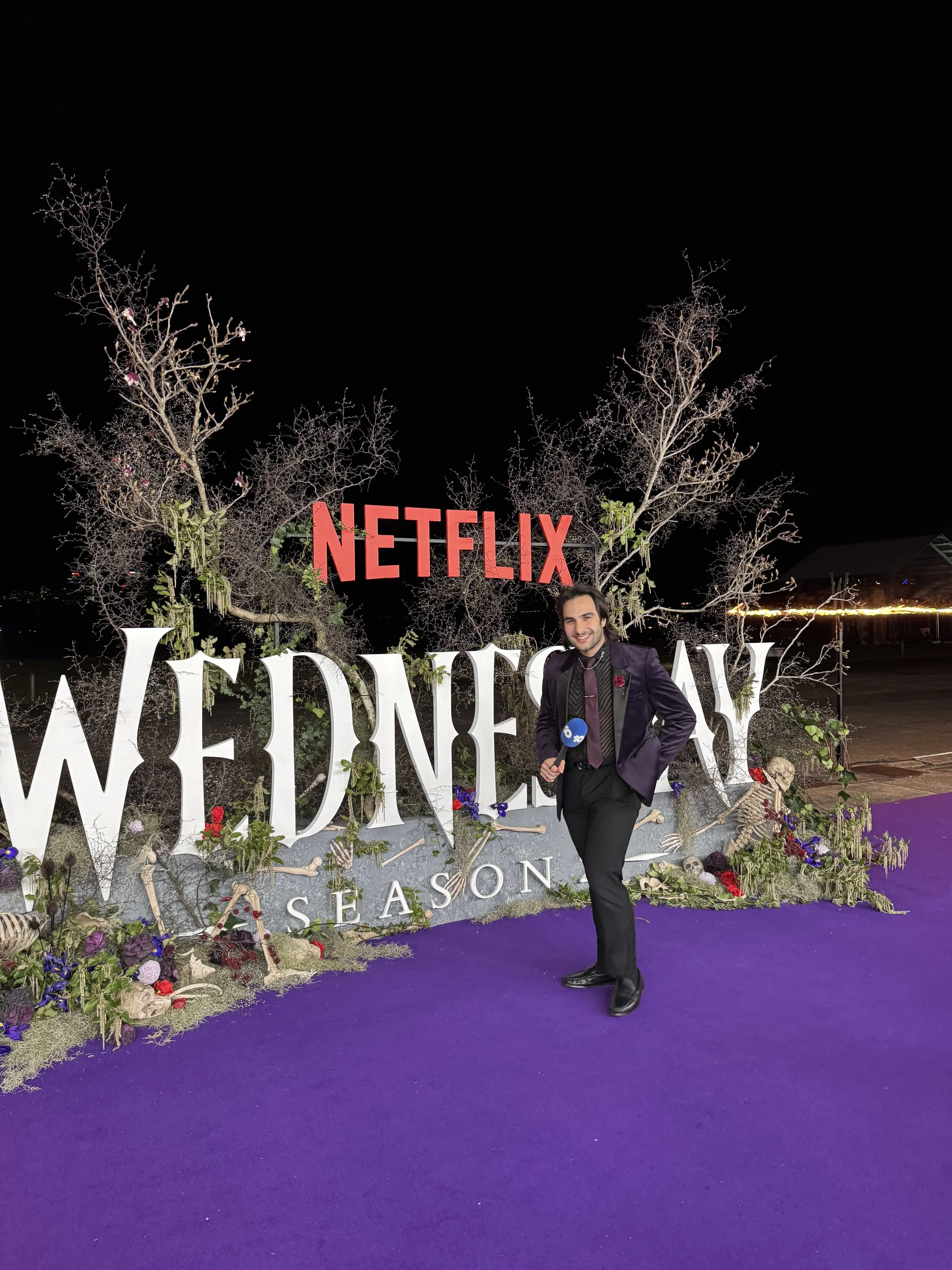 A person in a dark suit with a microphone stands in front of a wedding-themed backdrop featuring leafless trees, bones, flowers, and the words 'WEDDING SEASON' at night.