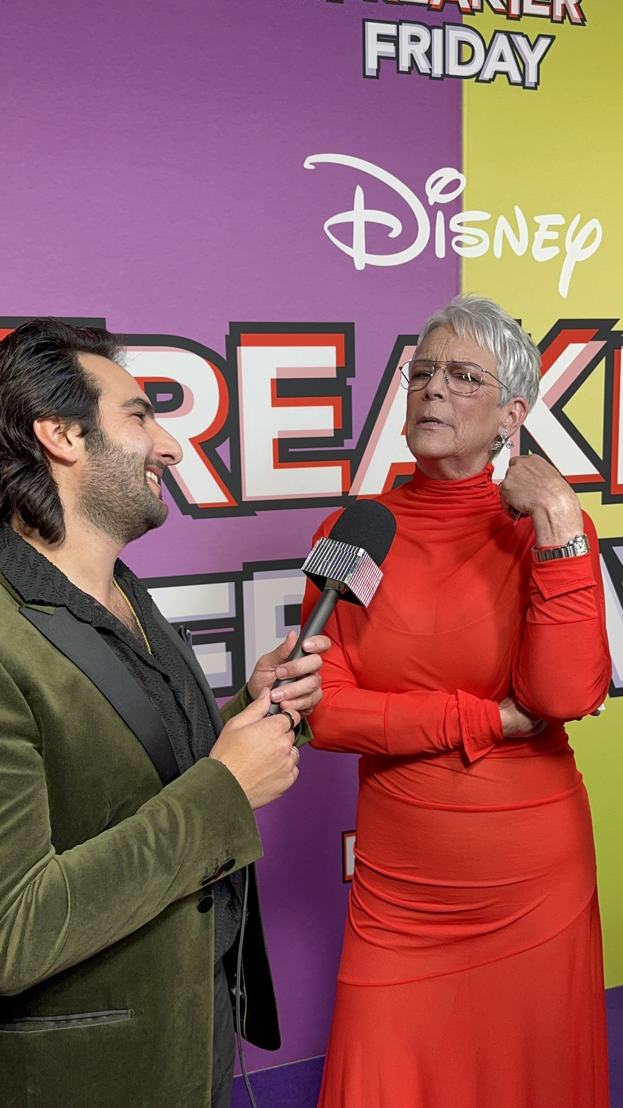 A woman in a bright red dress is being interviewed by a man holding a microphone at a Disney event. The background features a purple and yellow design with Disney and Marvel branding, including the words "February" and "Friday." The woman is adjustin