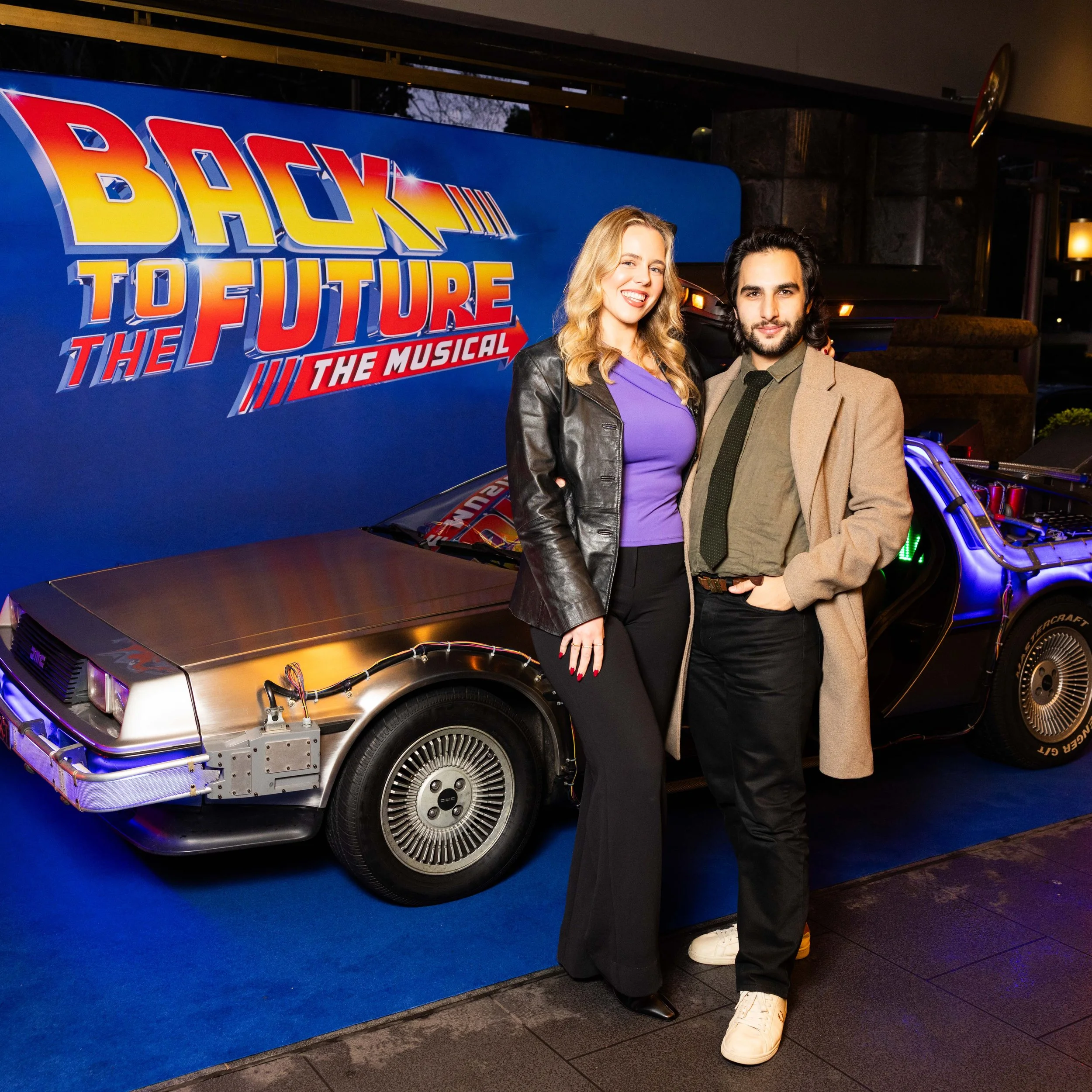 Two people, a woman with blonde hair and a man with dark hair and a beard, standing in front of a blue backdrop with the 'Back to the Future: The Musical' logo. The woman is smiling and wearing a black leather jacket and purple top, while the man is 