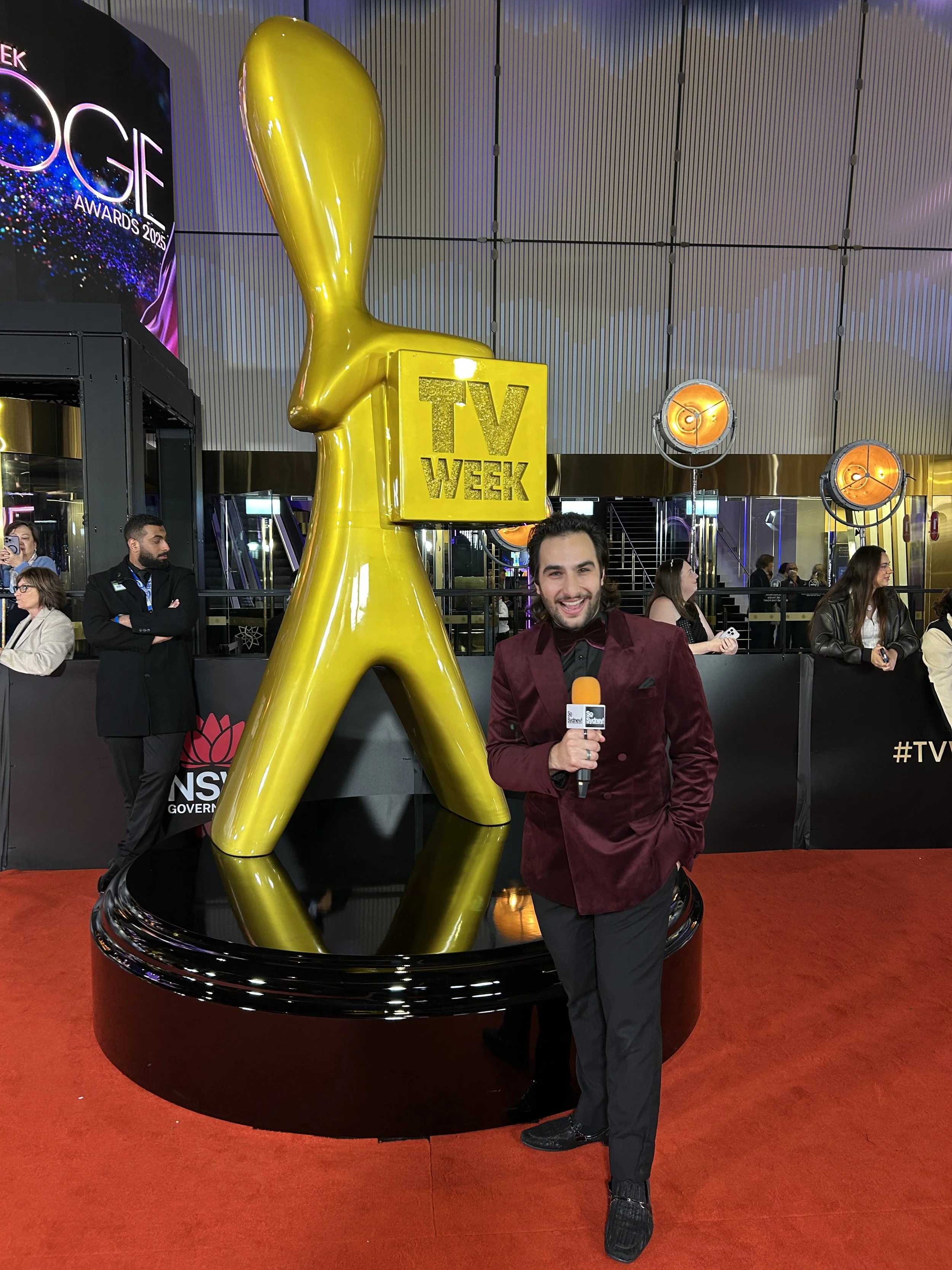 A man in a burgundy velvet blazer and black pants poses with a microphone at a red carpet event. Behind him is a large, gold, abstract TV award sculpture that says 'TV WEEK.' There are people in the background attending the event.