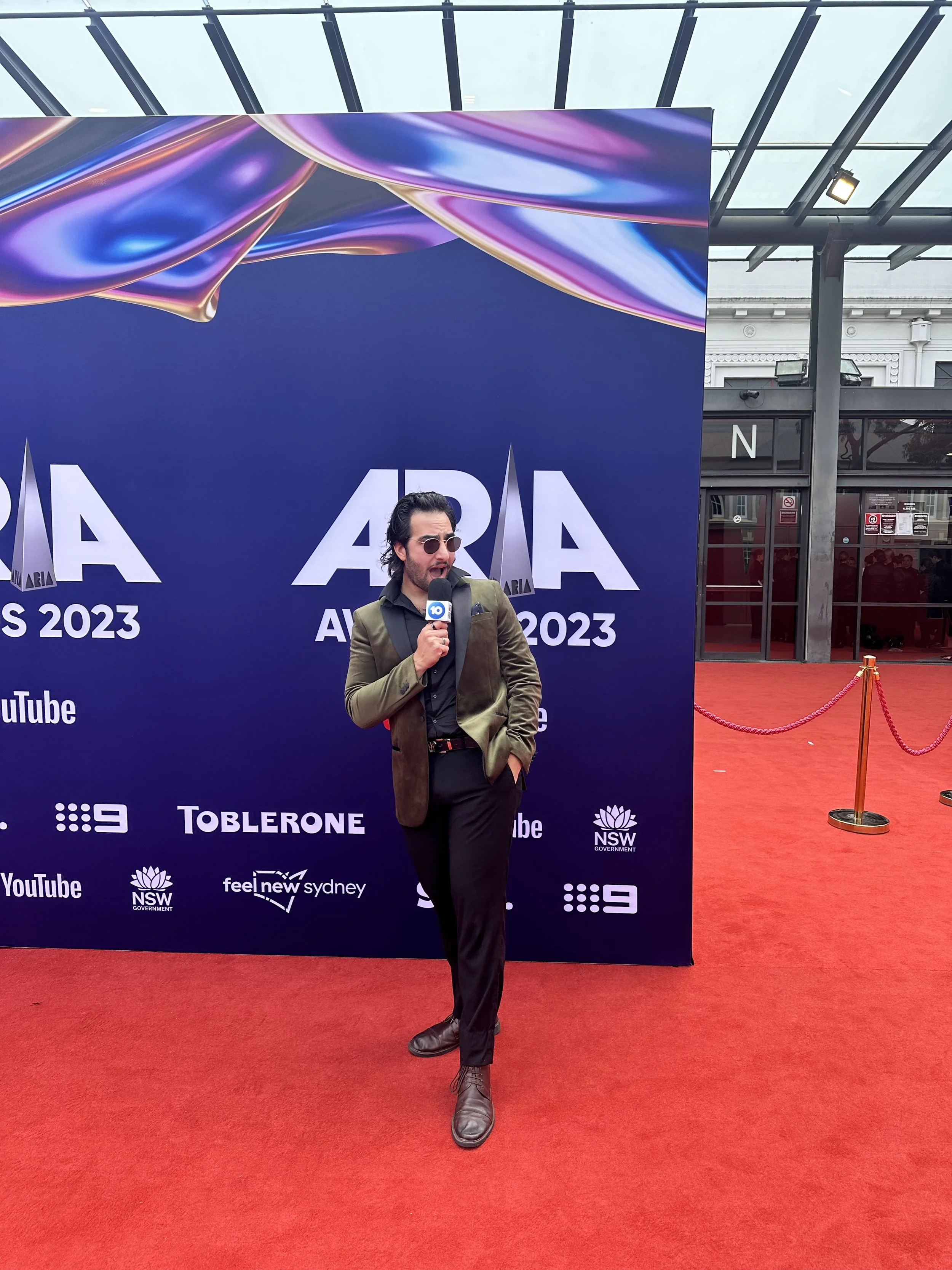 A man in a brown blazer and black pants stands on a red carpet, holding a microphone, in front of a backdrop with logos including YouTube, NSW Government, Feel New Sydney, and Toblerone, during the 2023 ARIA Awards.