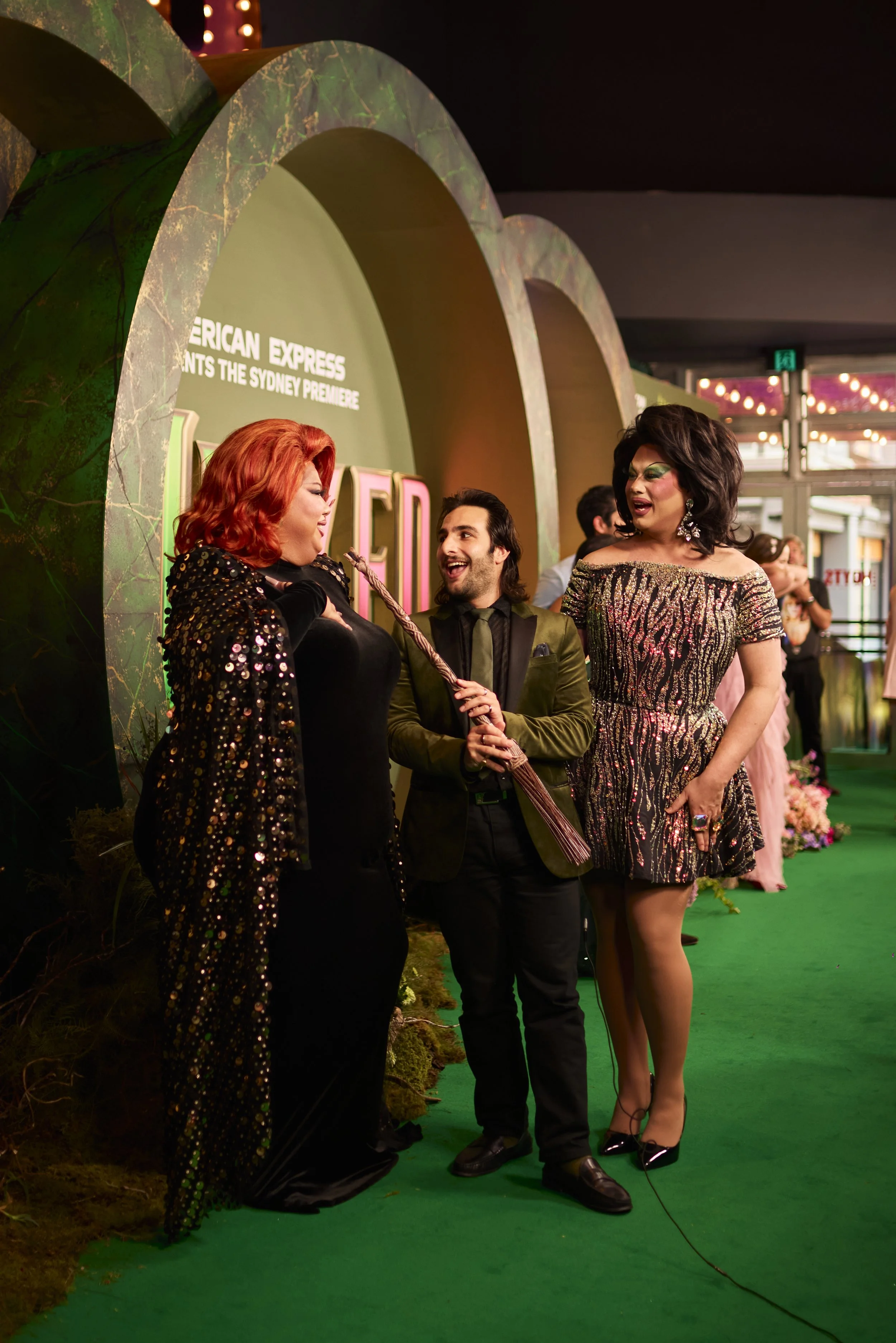 People dressed in glamorous attire at a red carpet event with a green backdrop and carpet.