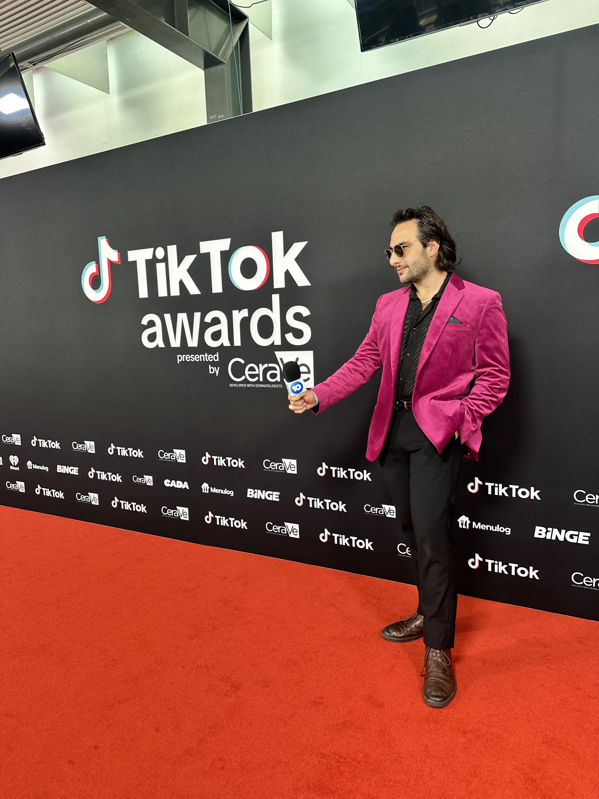 Man wearing pink blazer, black shirt, black pants, and sunglasses standing on red carpet at TikTok awards event, with TikTok and sponsors' logos on black backdrop.